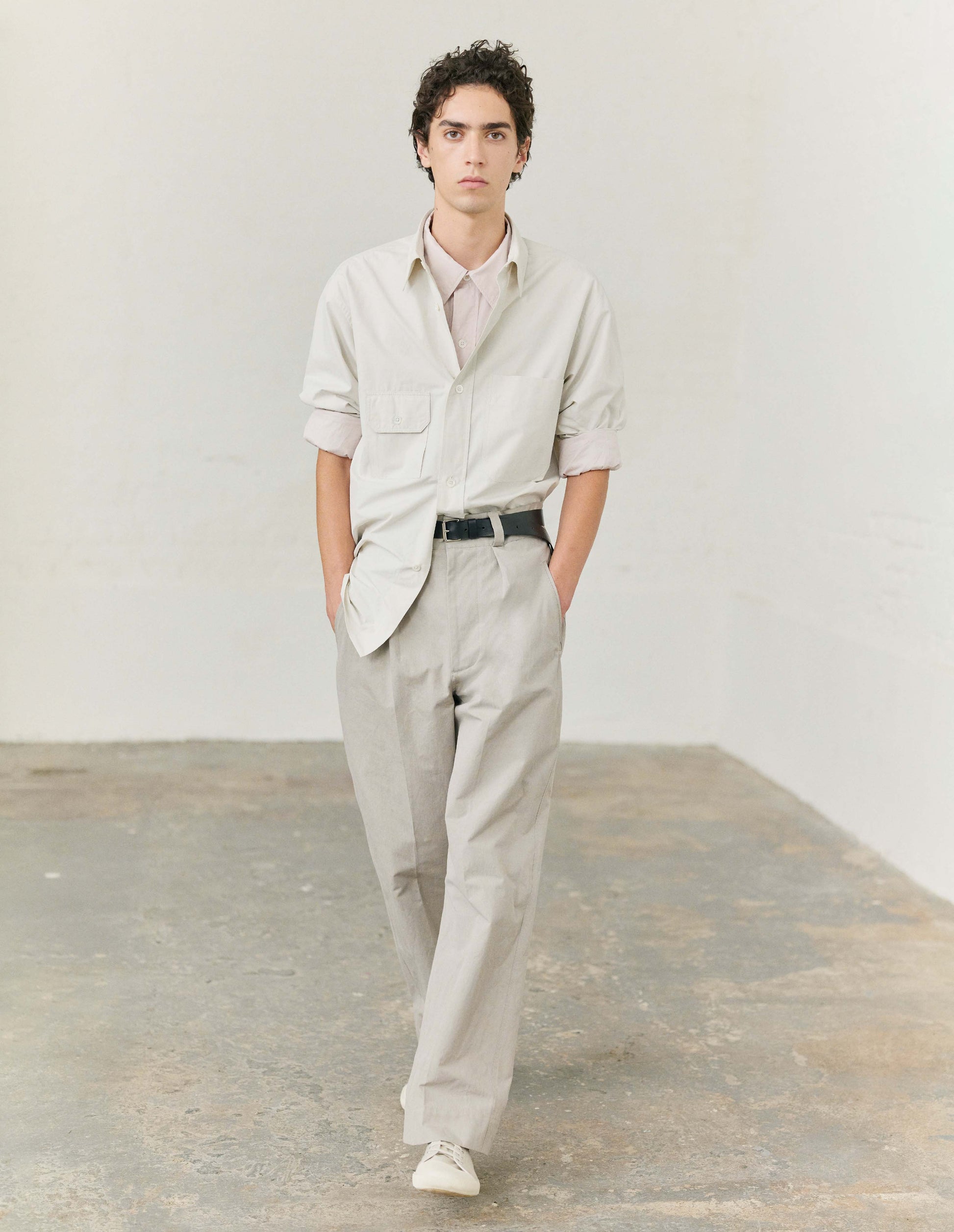 Full body image, front view of model wearing off white long sleeve cotton shirt with patch pocket and button-through flap pocket on chest and raised placket with mother of pearl buttons. Styled over a pink shirt and tucked into beige trousers, with black leather belt and off white canvas trainers. 