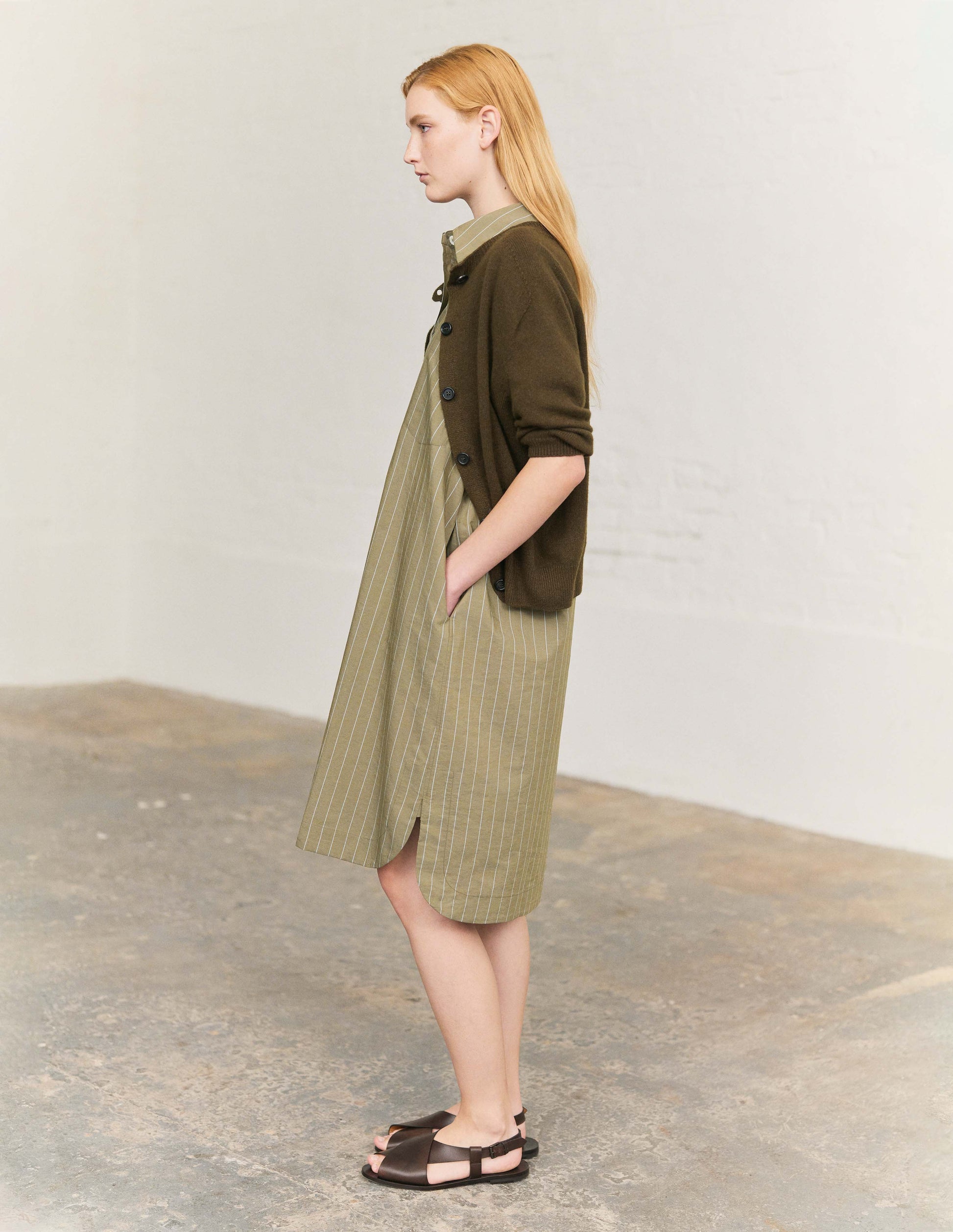 Full body image, front view of model wearing dark green cashmere cotton cardigan, with elbow length saddle sleeve and large horn buttons. Styled with light green and white vertical striped shirt dress and dark brown leather sandals. 