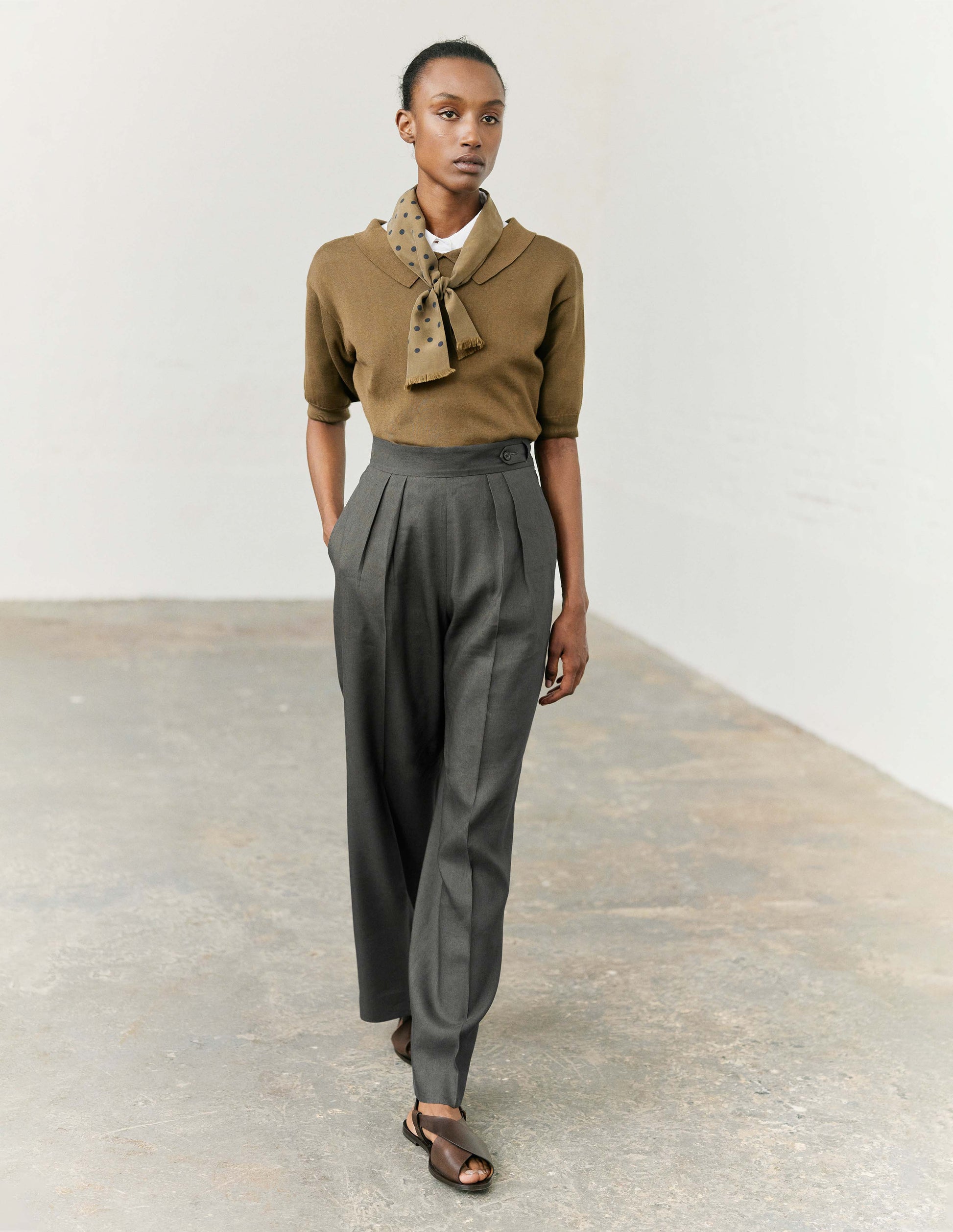 Full body image, front view of model wearing regular fit green split collar knitted t-shirt with ribbed cuffs and hem. Styled with white collared shirt, grey trousers, green and blue polka dot neckerchief and dark brown leather sandals.