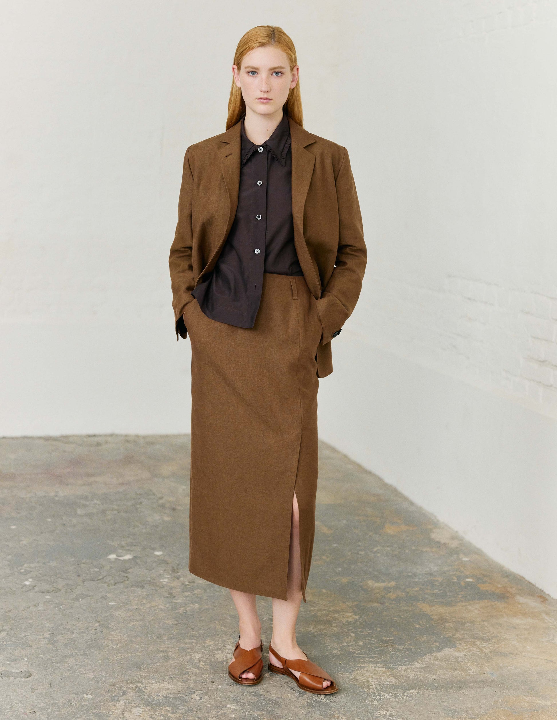 Full body image, front view of model wearing dark red cotton silk frilled collar long sleeve shirt, with mother of pearl button closure. Styled with brown blazer and matching brown skirt and light brown leather sandals.