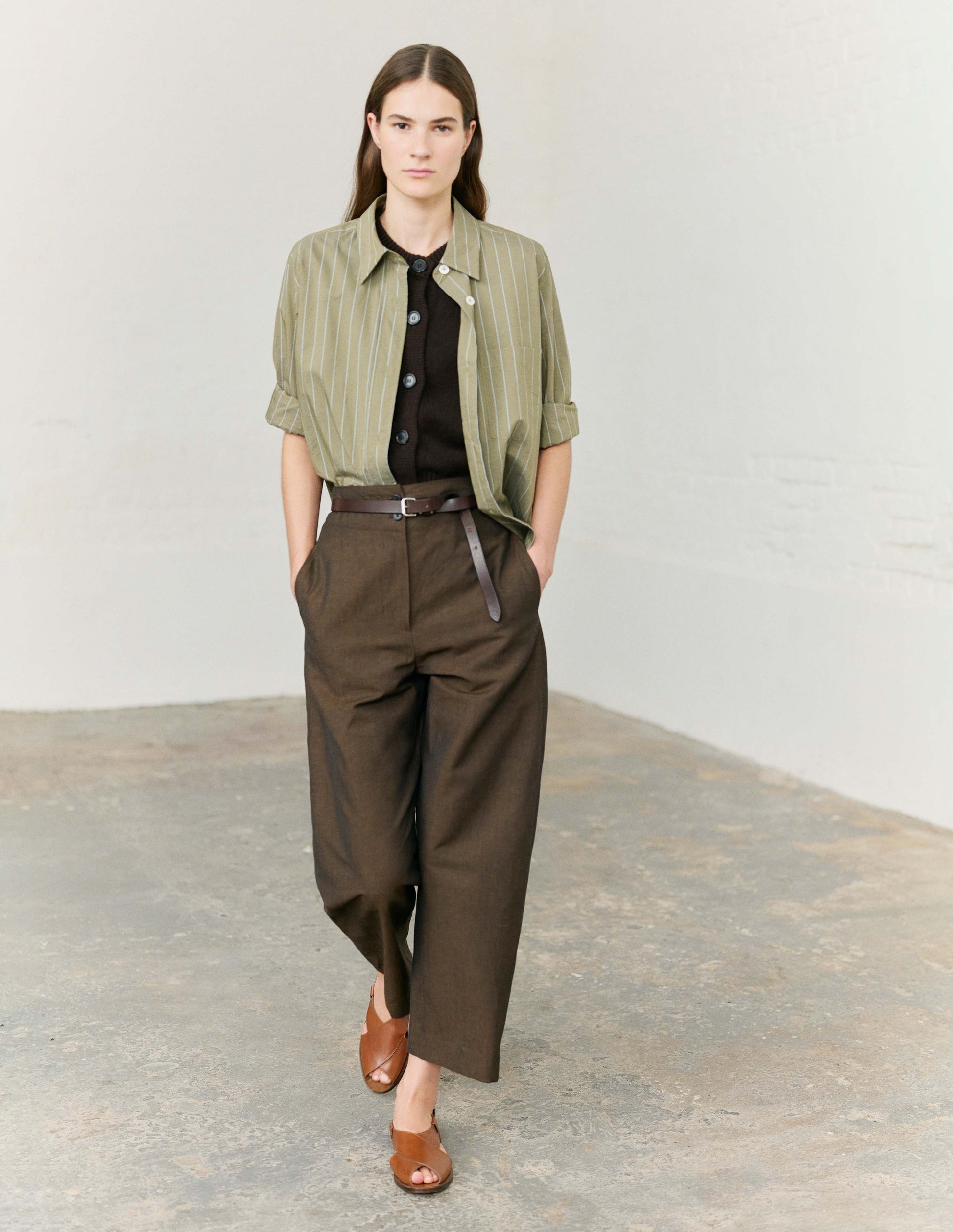 Full body image, front view of model wearing brown cotton linen trousers, with deep waistband, double horn button closure and slant side pockets. Styled with pale green and white vertical striped shirt, dark brown button up knitted vest, dark brown leather belt and light brown leather sandals. 
