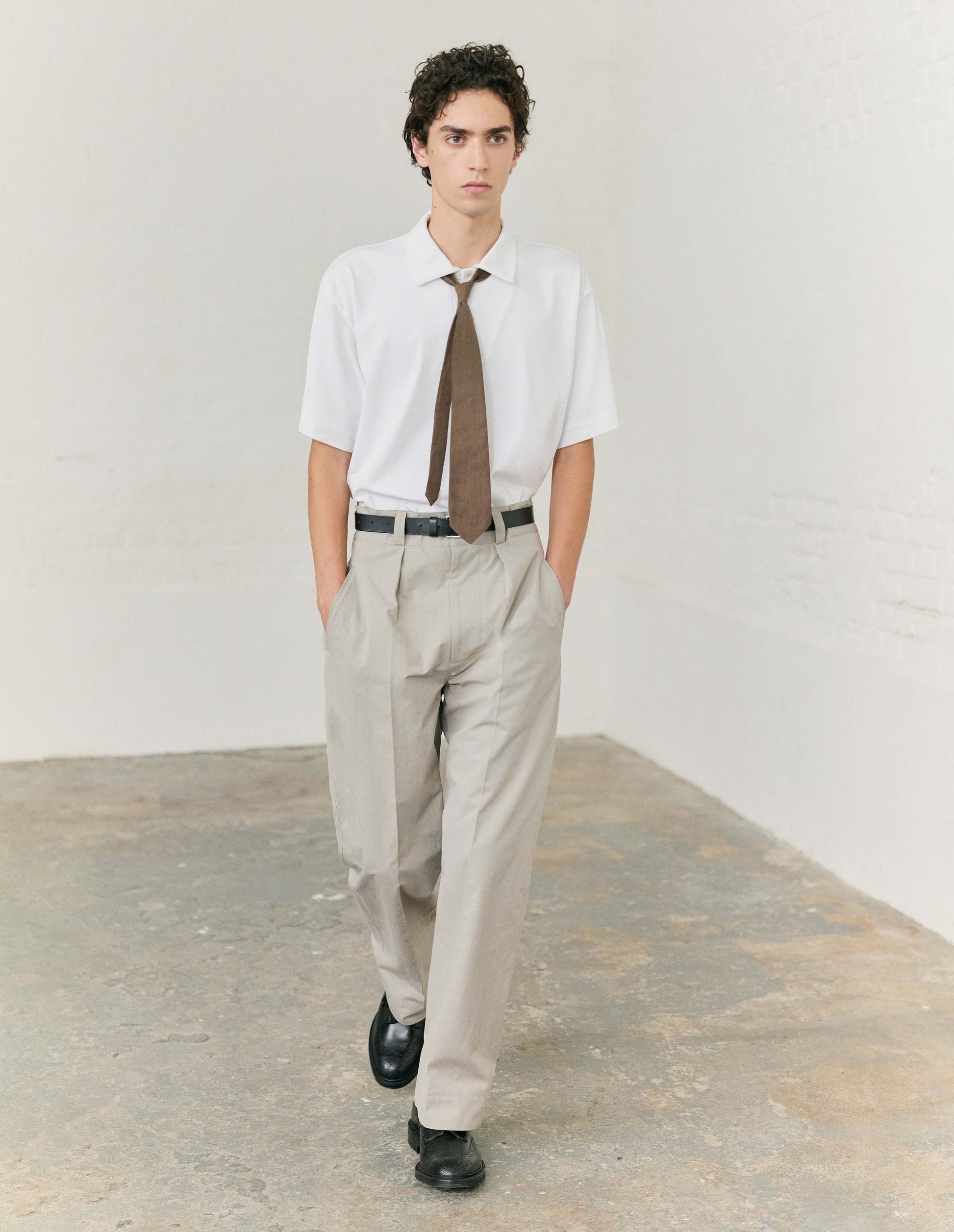 Full body image, front view of model wearing white short sleeve polo shirt with wide placket and horn buttons. Styled with a brown tie, tucked into beige trousers, with black leather belt and black leather shoes. 