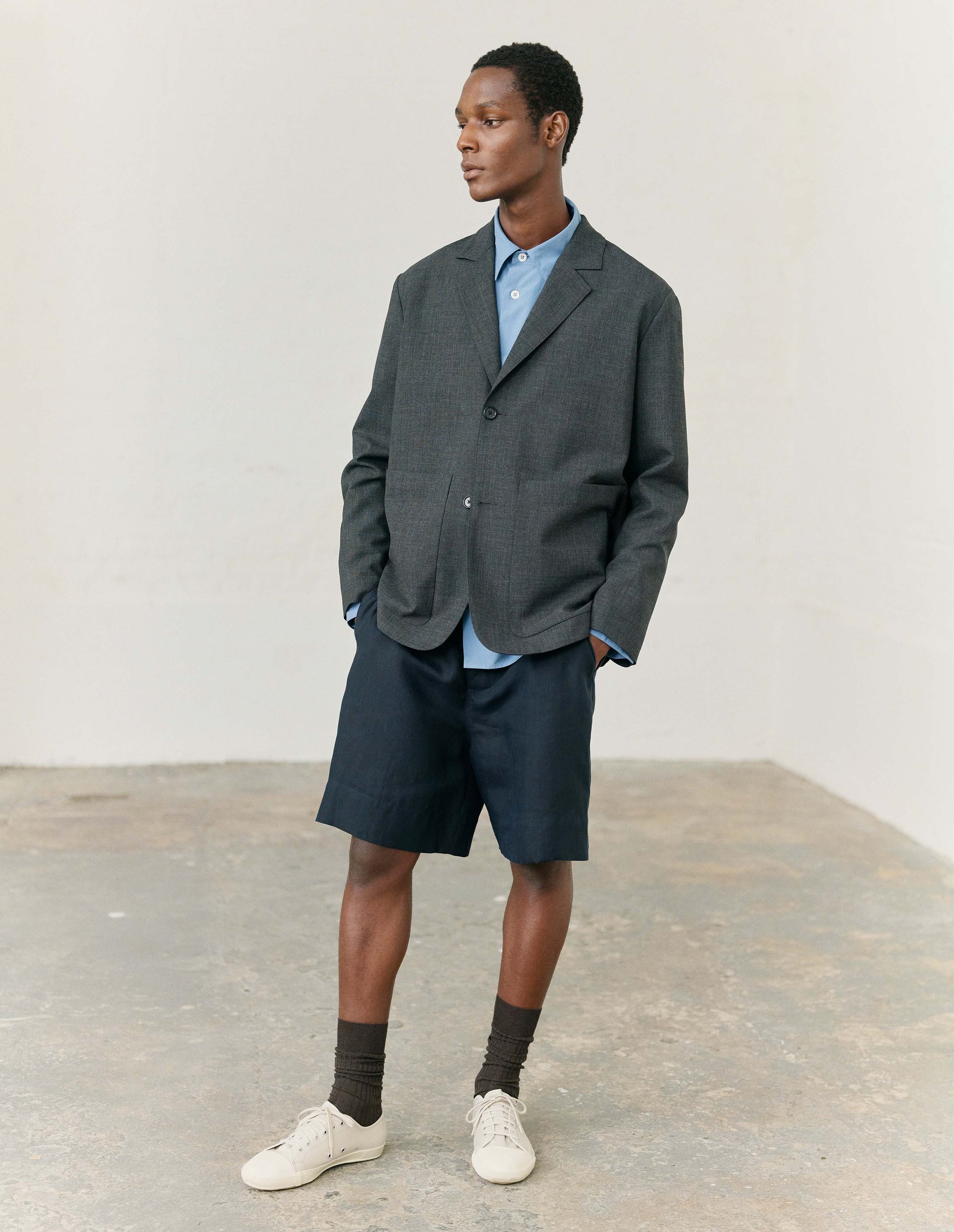Full body image, front view of model wearing blue cotton long sleeve shirt, with mother of pearl buttons, curved hem and chest patch pocket. Styled with dark navy blue shorts, black socks and black leather brogues. Styled with dark grey wool blazer, dark navy blue shorts, black socks and white canvas trainers.