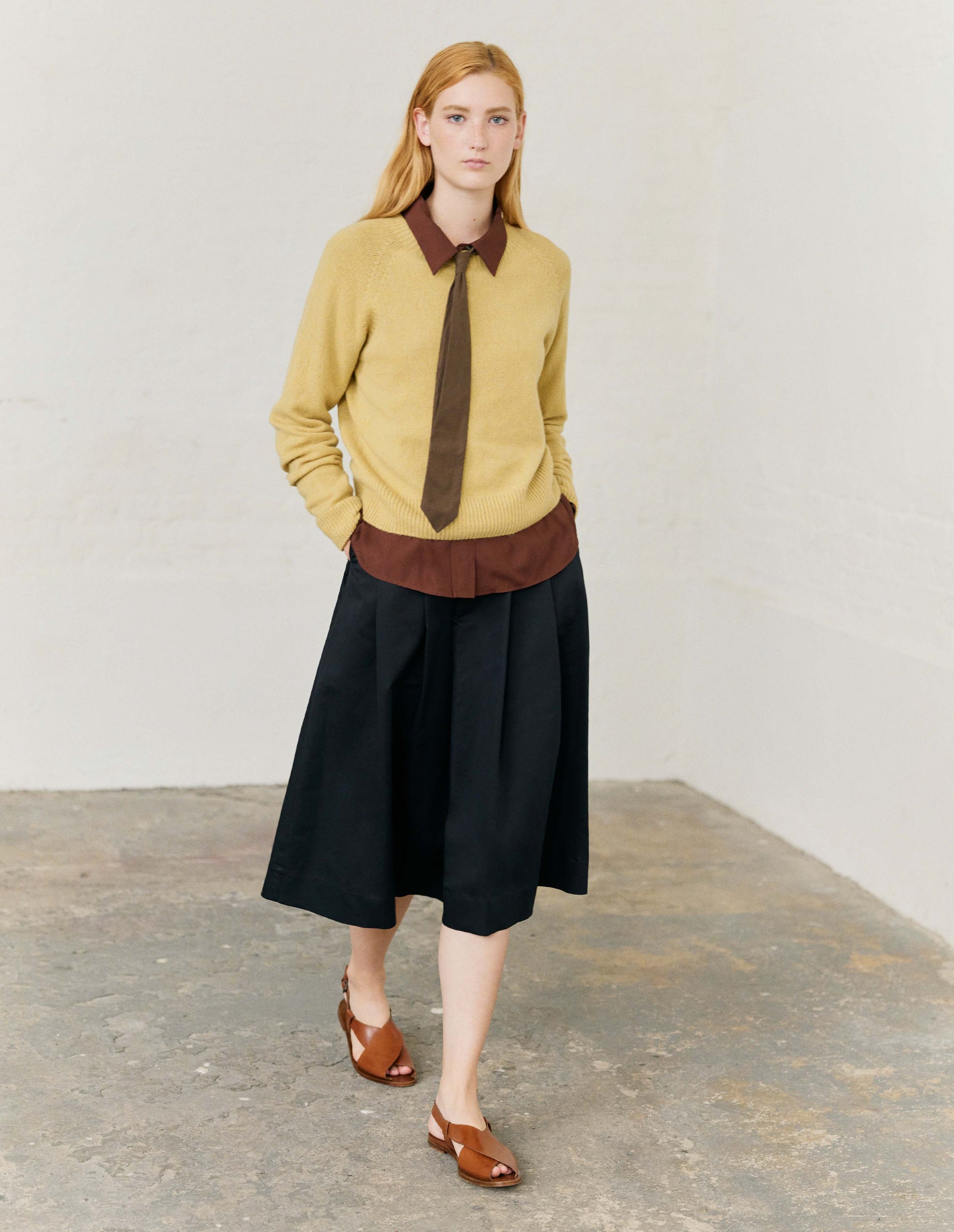 Full body image, front view of model wearing yellow merino cashmere long sleeve jumper, with raglan sleeve and contrast rib on neck, cuffs and hem. Styled with brown collared shirt, brown tie and light brown leather sandals.