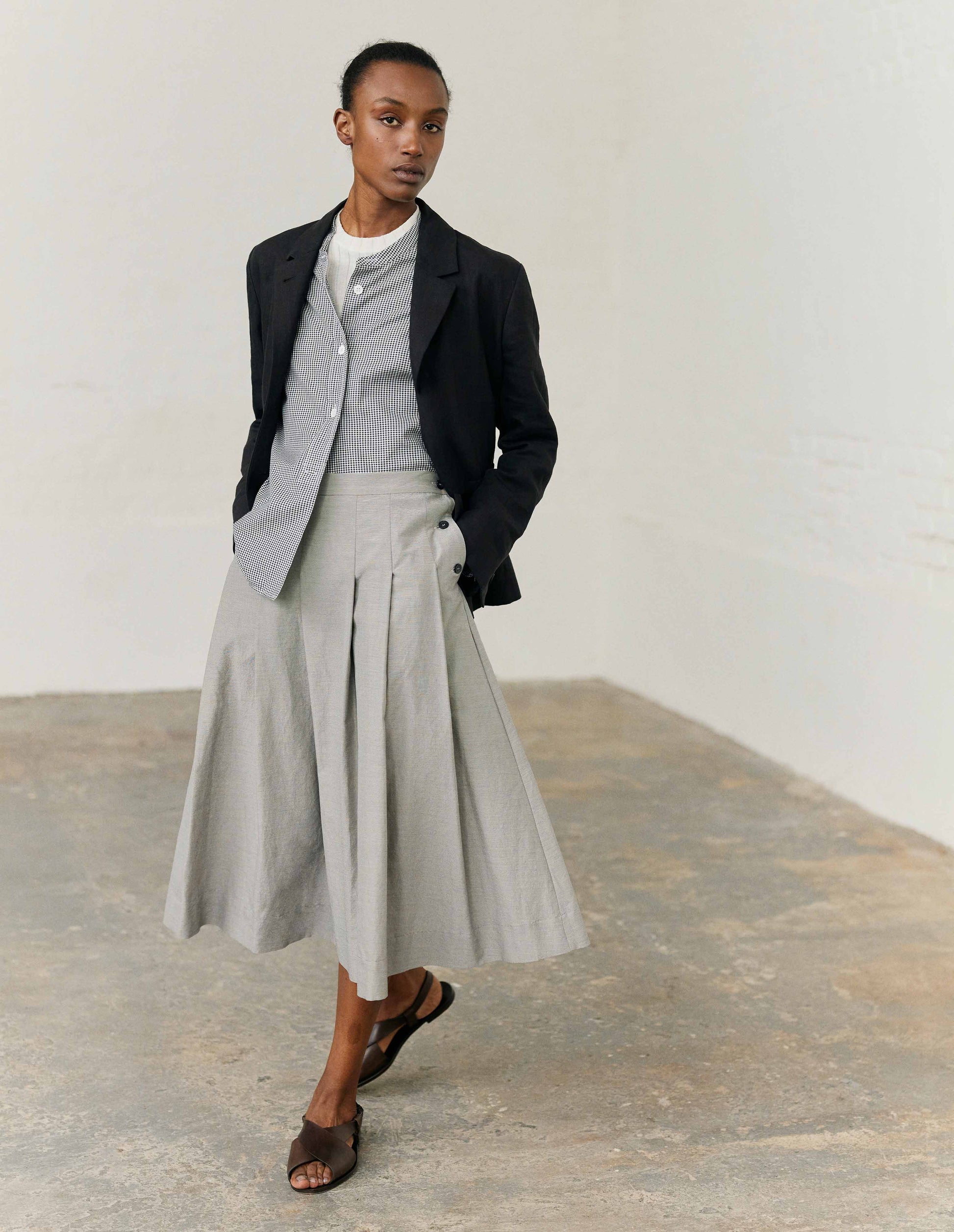 Full body image, front view of model wearing black linen blazer with horn button fastening lapel. Styled with white t shirt, black and white checked collarless shirt, light grey pleated skirt and dark brown leather sandals.