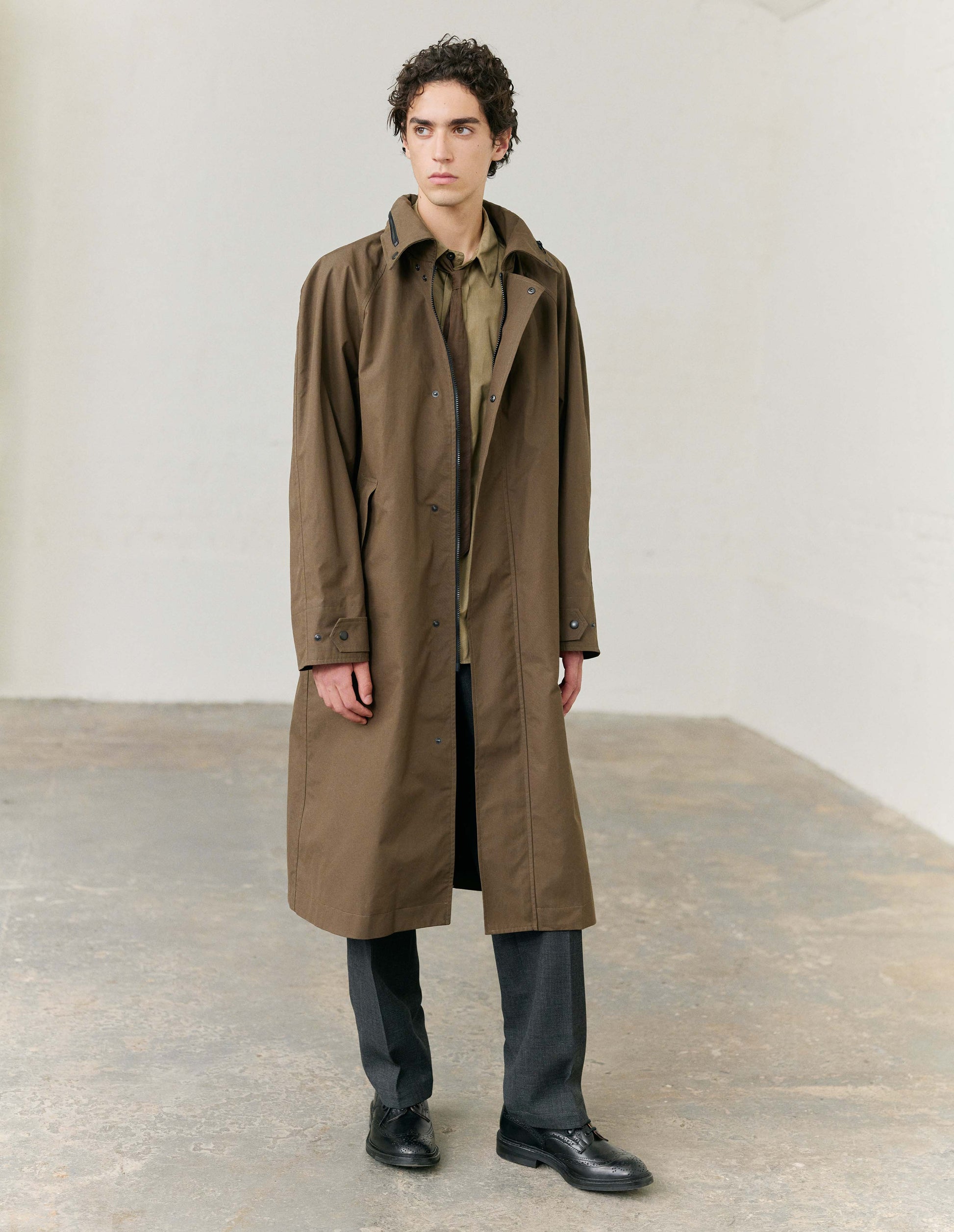 Full body image, front view of model wearing greenish brown ventile cotton poplin longline mac, with slant pockets, vislon zip and adjustable popper cuffs. Styled with light greenish brown shirt, brown tie, dark grey trousers and black leather shoes. 