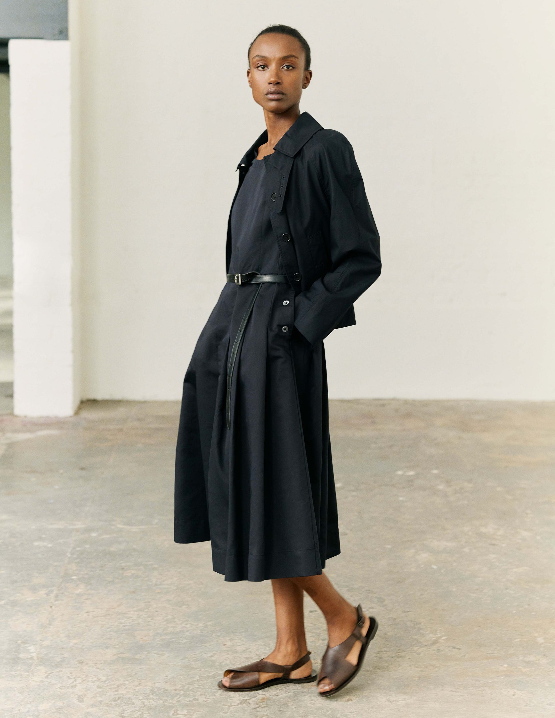 Full body image, front view of modelw wearing black ventile cotton poplin long sleeve short trench, with horn button closure, slant side pockets, adjustable storm tab and hanger loop on collar. Styled with black dress and black leather belt with dark brown leather sandals. 
