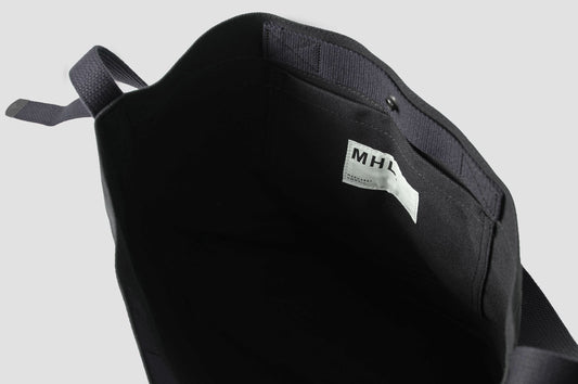 Black heavy cotton canvas shoulder tote bag with adjustable contrast nylon shoulder strap, interior patch pocket with white contrasting MHL. label attached and metal popper closure. Interior flat lay view.