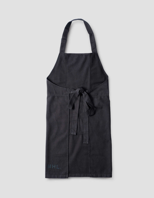 Black heavy cotton canvas apron with adjustable overhead strap, tie back straps and black MHL. stamped logo on right hand side. Back flat lay view.