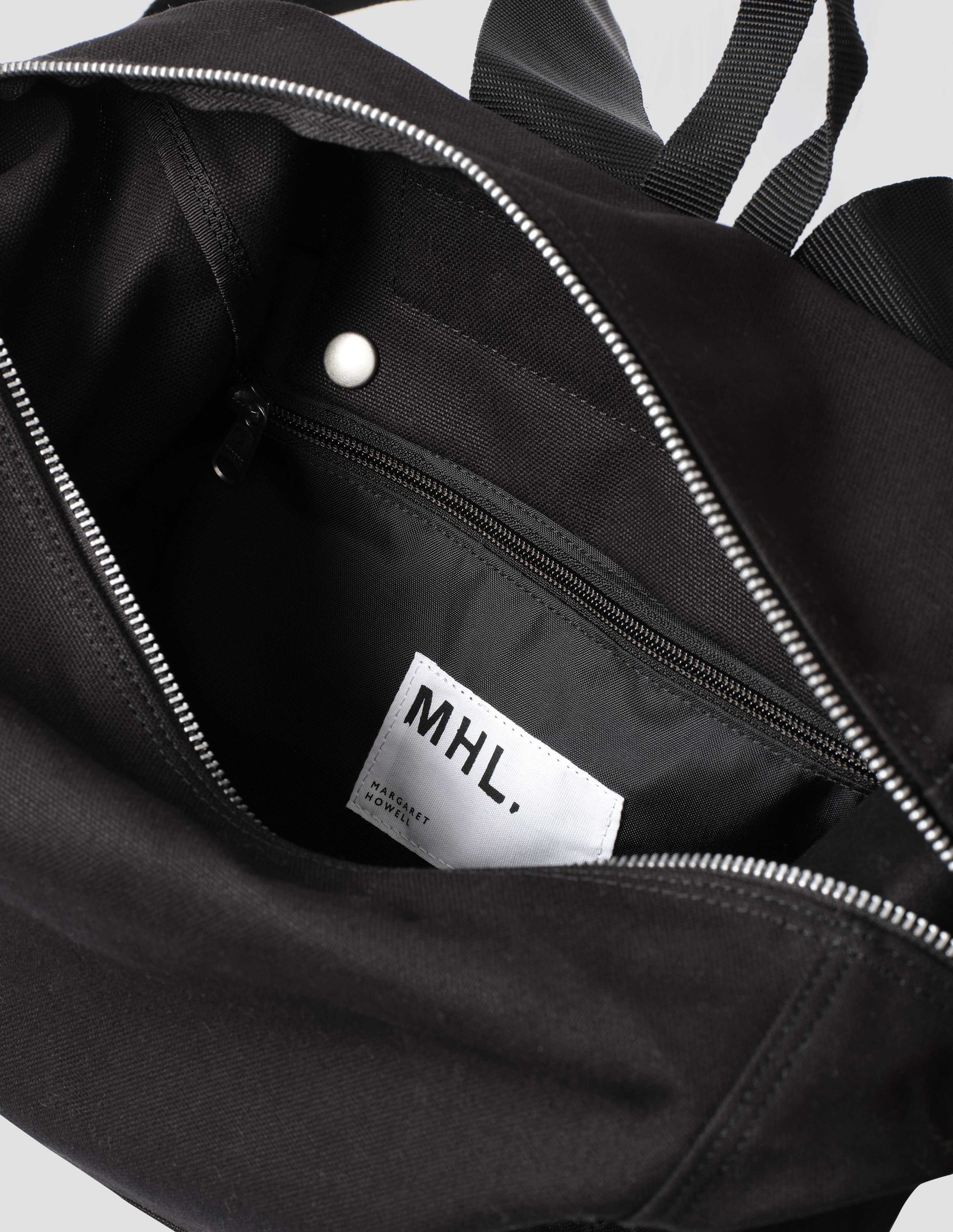 Black cotton canvas rucksack with contrast cotton webbing carry handles and zip fastening. Close up detail flat lay.