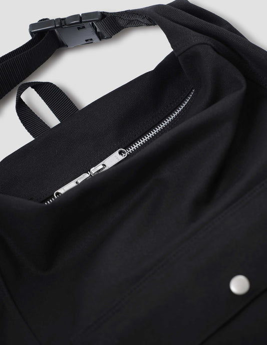 Black cotton canvas rucksack with contrast cotton webbing carry handles, adjustable clip, zip fastening and front pocket with popper closure. Close up detail flat lay.