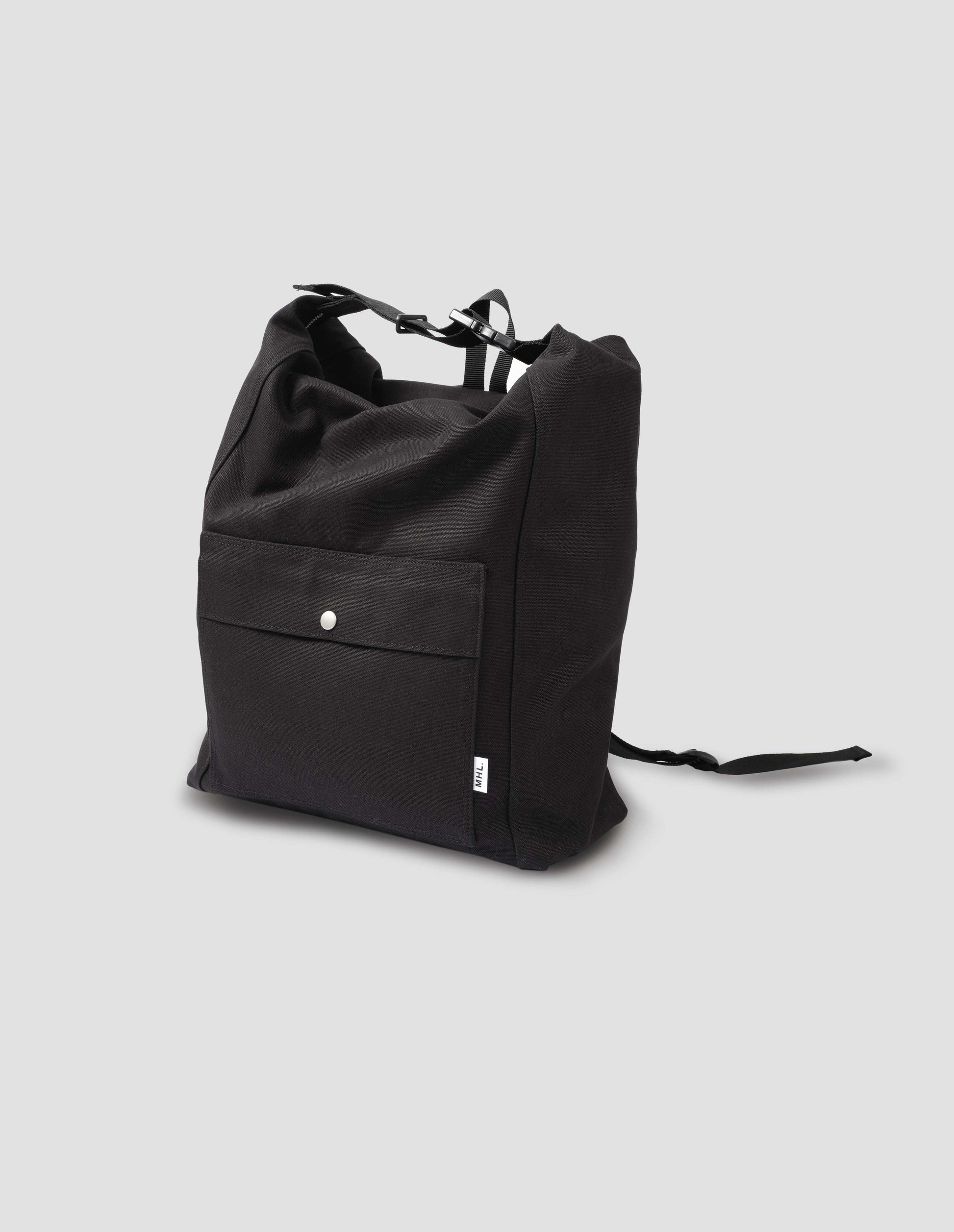 Black cotton canvas rucksack with contrast cotton webbing carry handles, adjustable clip, zip fastening, front pocket with popper closure, and white tag with MHL. logo. Front flat lay view.