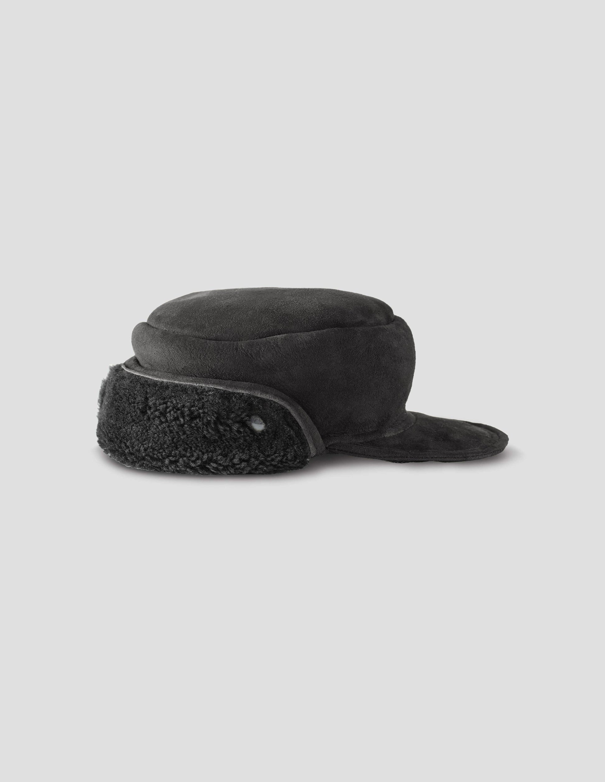 Black shearling press stud cap with peak and black curly merino sheepskin earflaps. Side flat lay view.