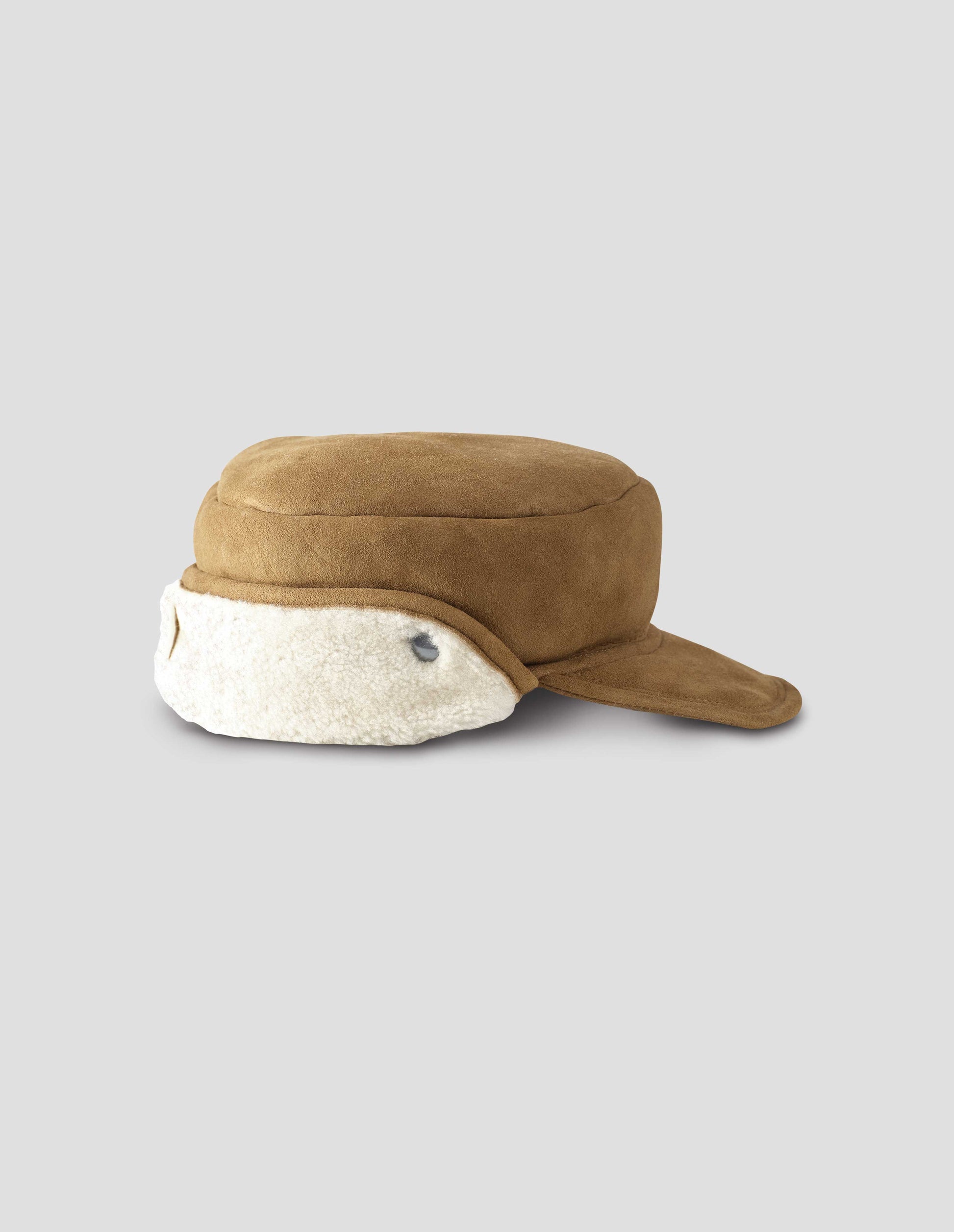 Light brown shearling cap with peak and metal press studs on white curly merino sheepskin ear flaps. Side flat lay view.