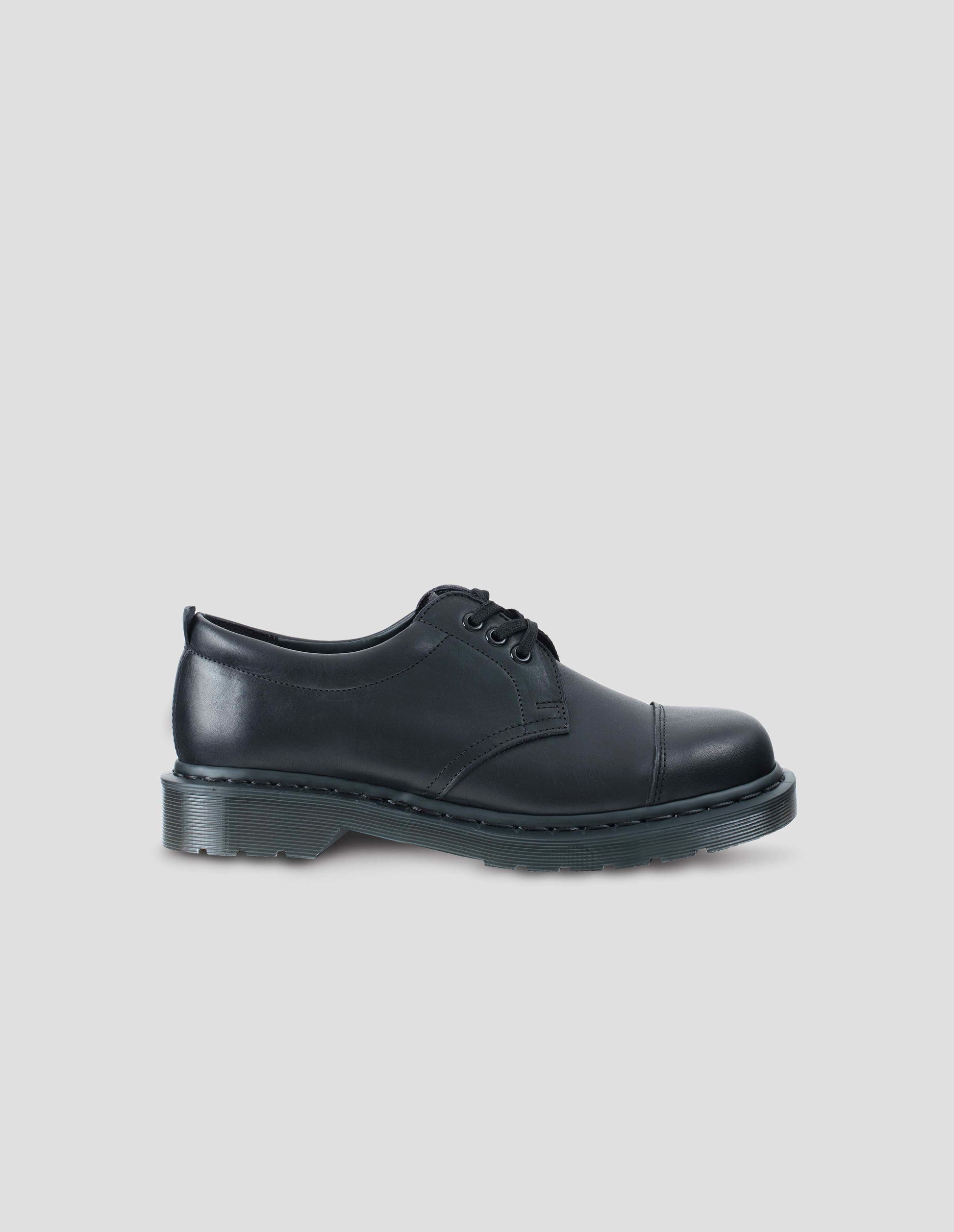 Single black matte grained leather 3-eyed shoe features flat cotton laces, double-stitched toe cap, padded heel collar. Dark grey Dr. Martens air-cushioned sole. Side view flat lay.
