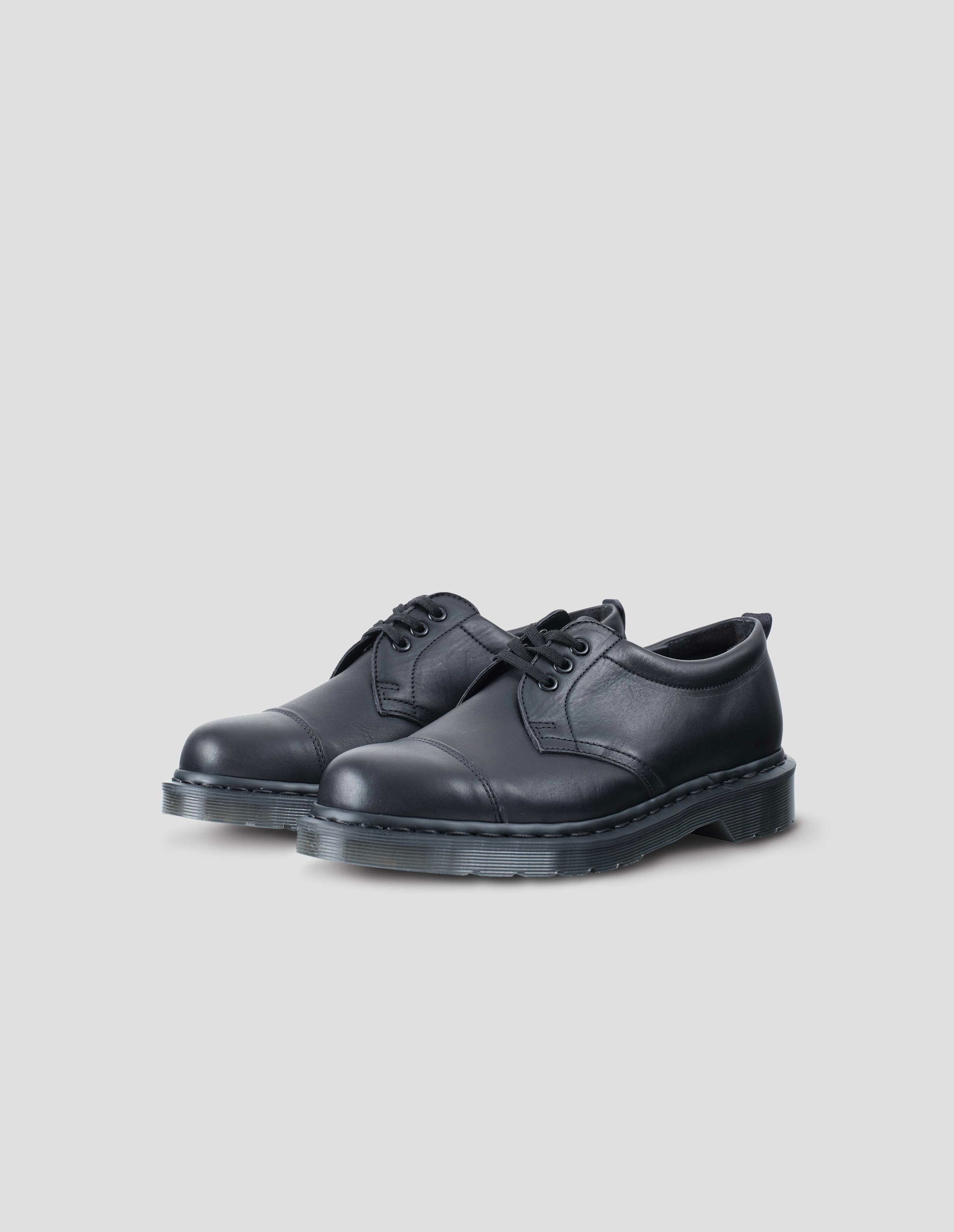 A pair of black matte grained leather 3-eyed shoe features flat cotton laces, double-stitched toe cap, padded heel collar. Dark grey Dr. Martens air-cushioned sole. Side flat lay view.