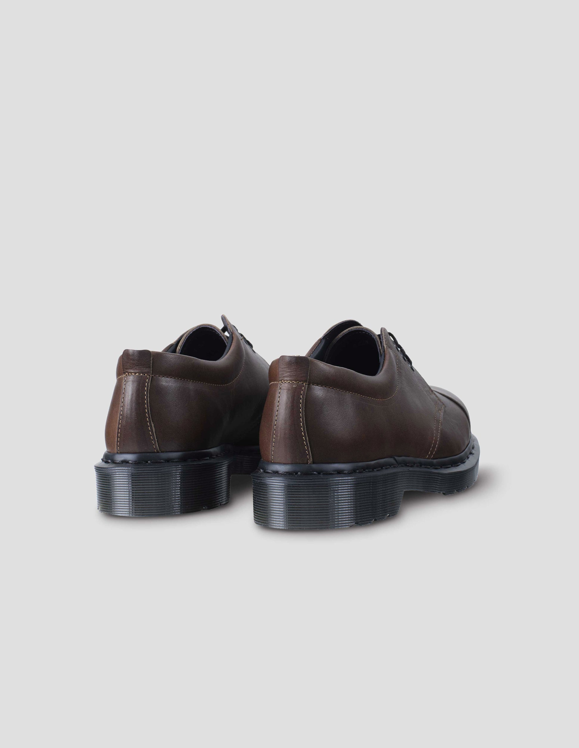 A pair of dark brown matte grained leather  shoe features a padded heel collar. Dark grey Dr. Martens air-cushioned sole. Back flat lay view.