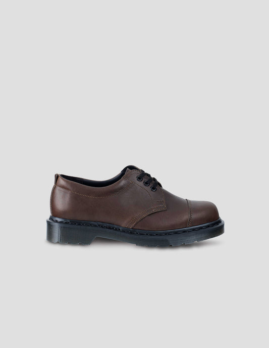 Single dark brown matte grained leather 3-eyed shoe features flat cotton laces, double-stitched toe cap, padded heel collar. Dark grey Dr. Martens air-cushioned sole. Side view flat lay.