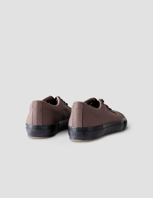 A pair of brown cotton canvas trainers with metal eyelet, black laces and contrast vulcanised rubber sole with MHL. logo stamped on back of sole. Back flat lay view.