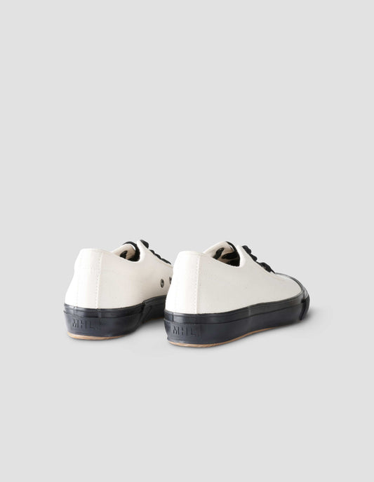 A pair of white cotton canvas trainers with metal eyelet, black laces and contrast vulcanised rubber sole with MHL. logo stamped on back of sole. Back flat lay view.