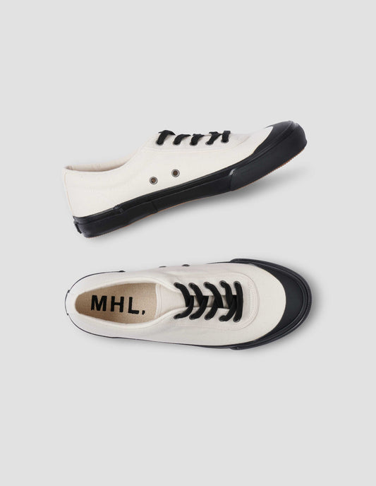 A pair of white cotton canvas trainers with metal eyelet, black laces and contrast vulcanised rubber sole. View from above flat lay view.