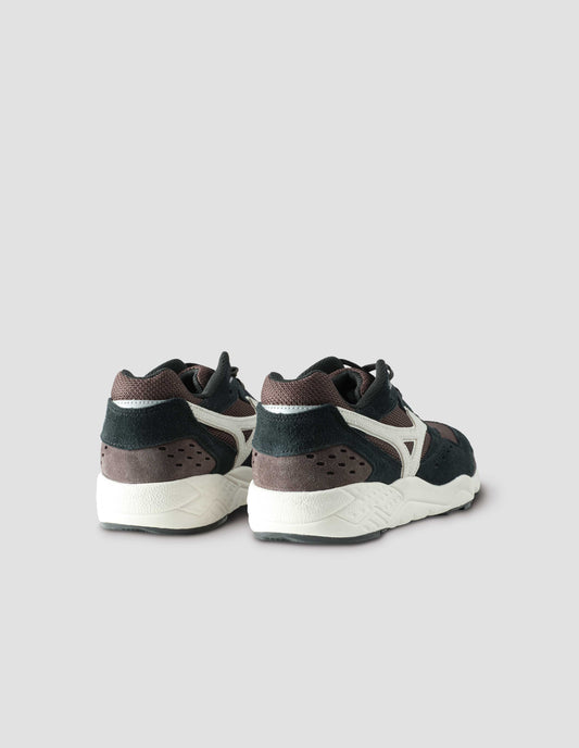 A pair of black and dark brown suede trainers with mesh insert and off white sole, with off white Mizuno logo running through the side. Back view flat lay.