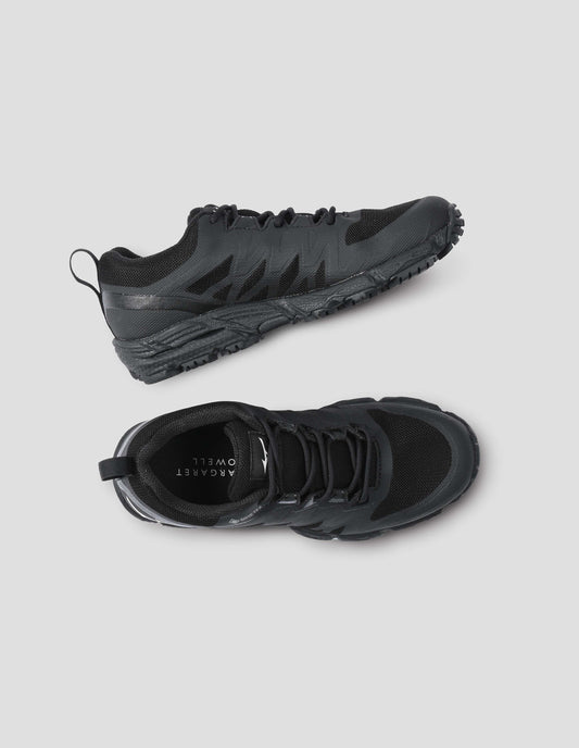 Black lace up trainers with rubber sole and fabric upper, pull loop on heel and  white Mizuno logo on tongue, white Margaret Howell logo on sole. Front and side flat lay view.