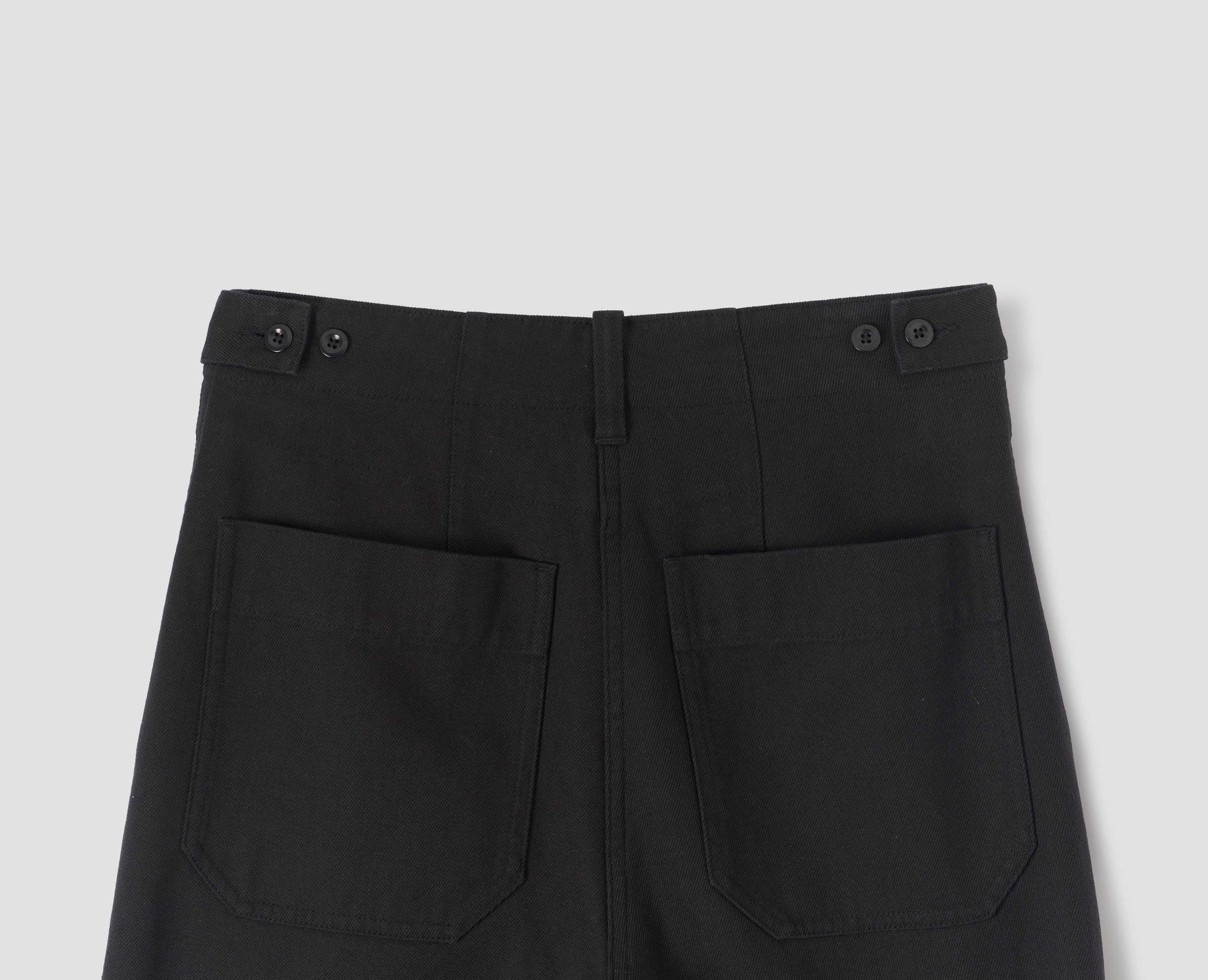 Black cotton wool trousers with button tab side adjusters, belt loop and patch back pockets. Back flat lay view.