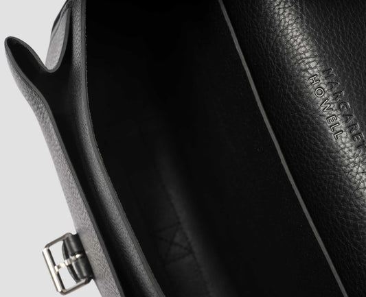 Black fine grain leather satchel bag with pleat expandable exterior pocket and interior slip pocket and embossed Margaret Howell logo detail. Fabric swatch.