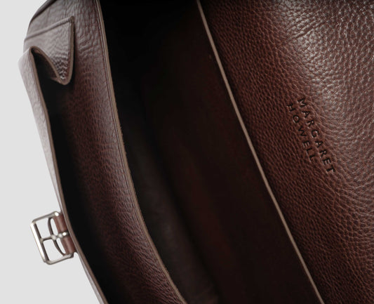 Brown fine grain leather satchel bag with pleat expandable exterior pocket and interior slip pocket and embossed Margaret Howell logo detail. Fabric swatch.