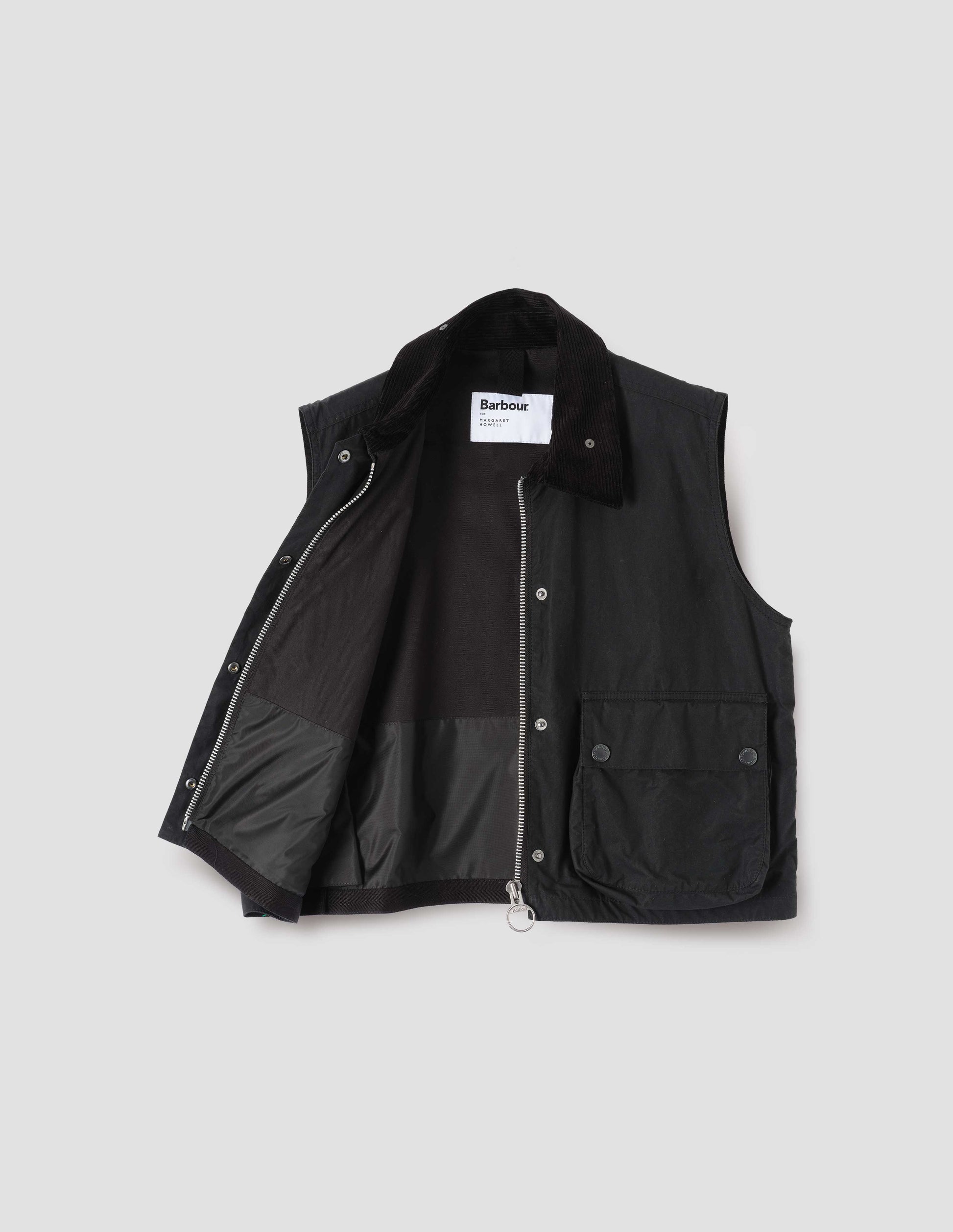 Black cotton drill lined boxy gilet with storm tab on corduroy collar, two-way zip fastening and press stud fastening storm flap, large bellows pockets with press stud fastening envelope closure. Open flat lay view.