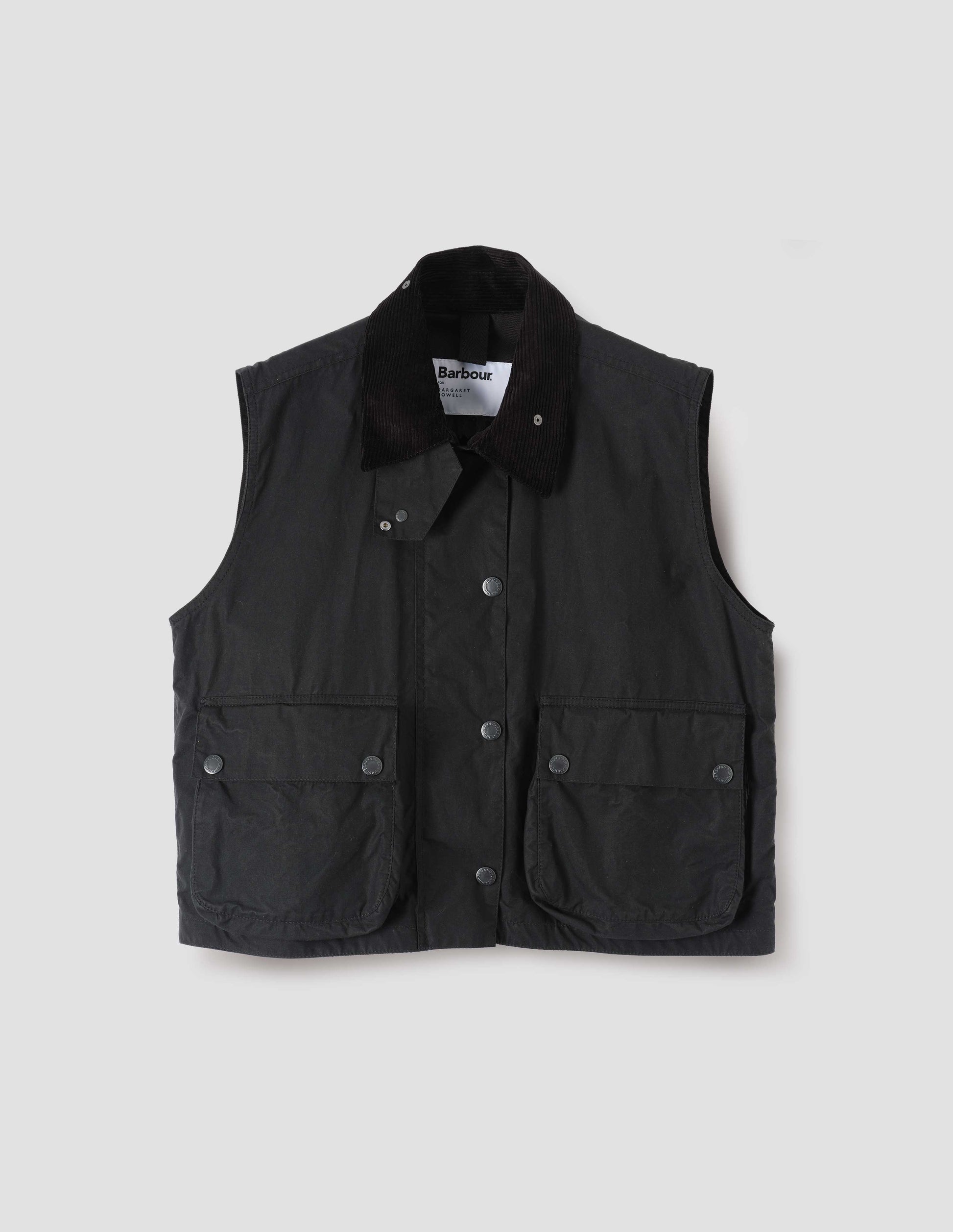 Black boxy gilet with storm tab on corduroy collar, press stud fastening storm flap, large bellows pockets with press stud fastening envelope closure. Front flat lay view.