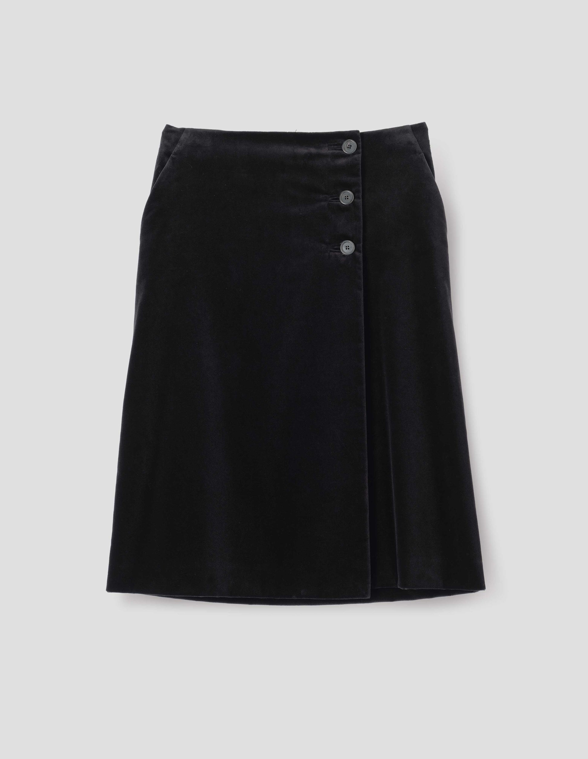 Black velvet a-line skirt with deep wrap, horn button fastening and slant side pockets. Front flat lay view.