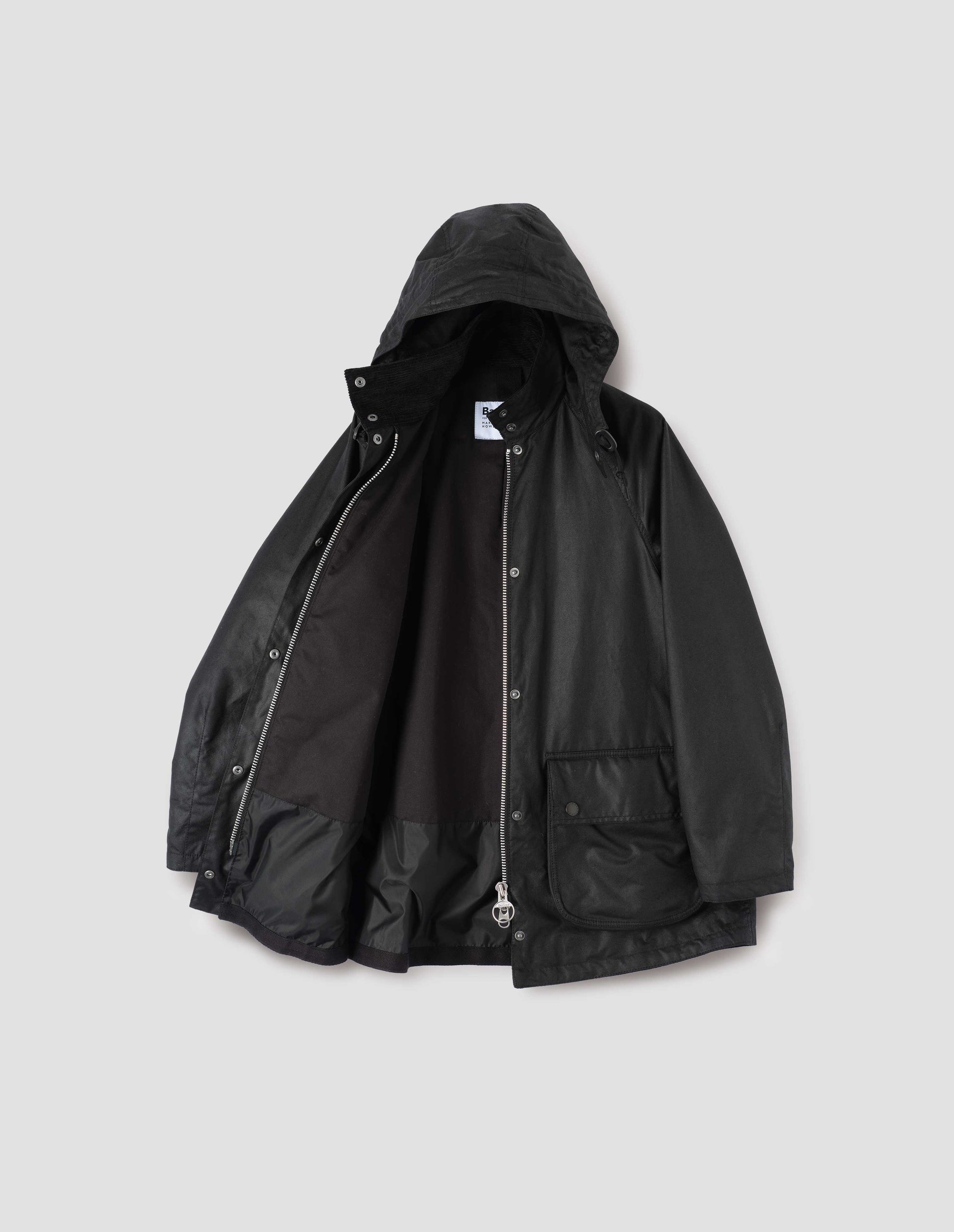 Black cotton drill lined parka with stand collar and hood with wire adjustable peak, raglan sleeve, two way zip fastening, press stud fastening storm flap and large bellows pockets with press stud fastening envelope closure. Open flat lay view.
