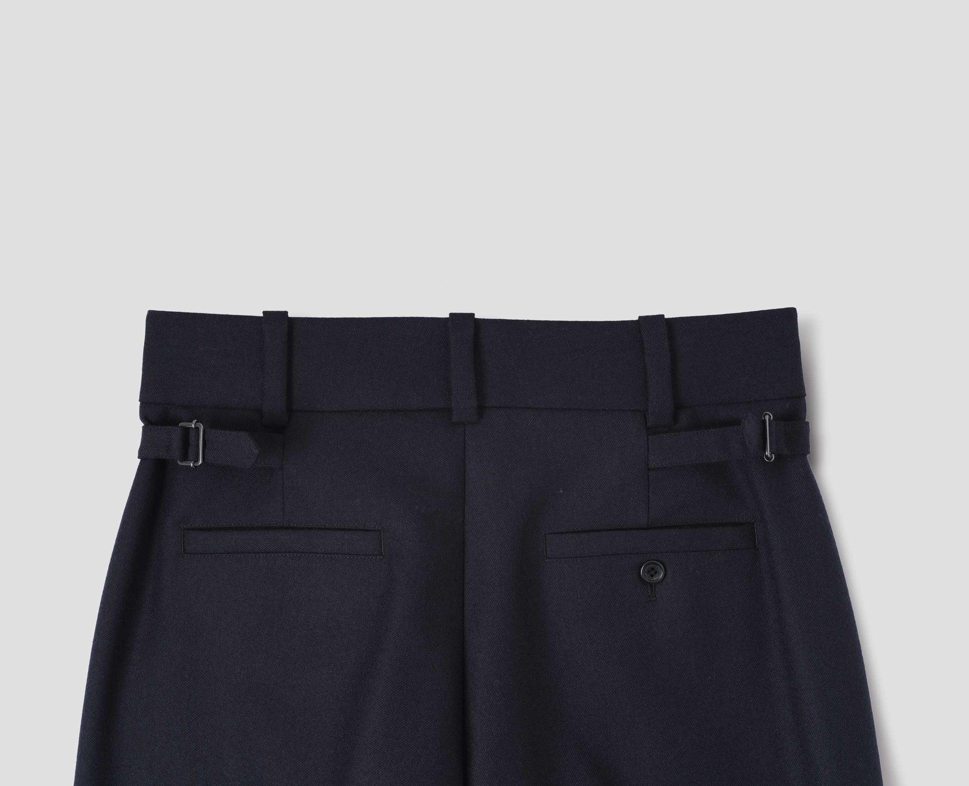 Dark blue wool trousers with belt loops on wide waistband, side adjusters and two welt back pockets, one with horn button fastening. Back flat lay view.