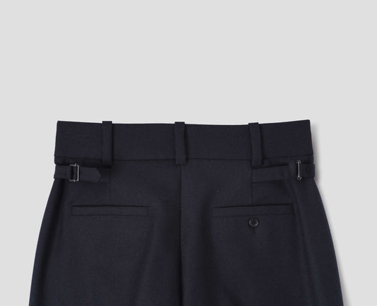 Dark blue wool trousers with belt loops on wide waistband, side adjusters and two welt back pockets, one with horn button fastening. Back flat lay view.