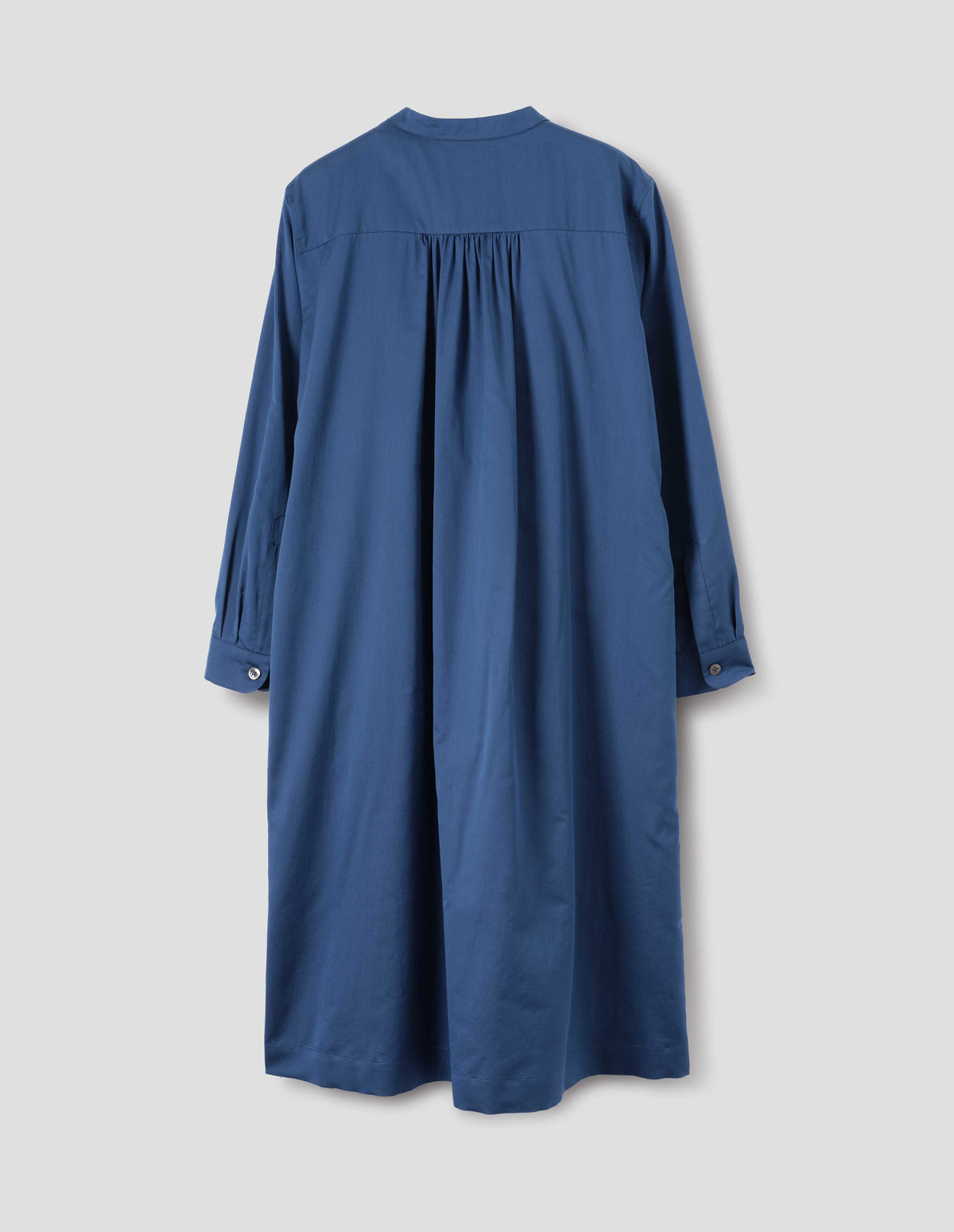 Blue cotton silk collarless dress with wide gathering at back yoke, shirt sleeves with mother of pearl buttons on narrow cuffs, side seam pockets and split hems. Back flat lay view.