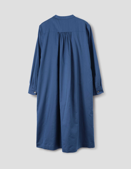 Blue cotton silk collarless dress with wide gathering at back yoke, shirt sleeves with mother of pearl buttons on narrow cuffs, side seam pockets and split hems. Back flat lay view.