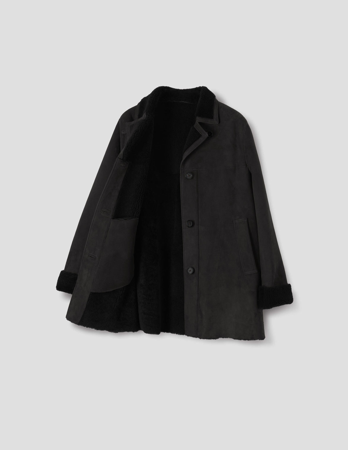 Black sheepskin coat with contrasting curly merino collar revere with narrow sueded binding, wide placket with button loop closure, horn button fastening, front yoke, slanted single welt front pockets, curly merino inside, and raw edges on contrasting curly merino turn-up cuffs and hem. Open flat lay view.