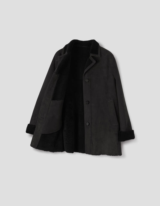 Black sheepskin coat with contrasting curly merino collar revere with narrow sueded binding, wide placket with button loop closure, horn button fastening, front yoke, slanted single welt front pockets, curly merino inside, and raw edges on contrasting curly merino turn-up cuffs and hem. Open flat lay view.