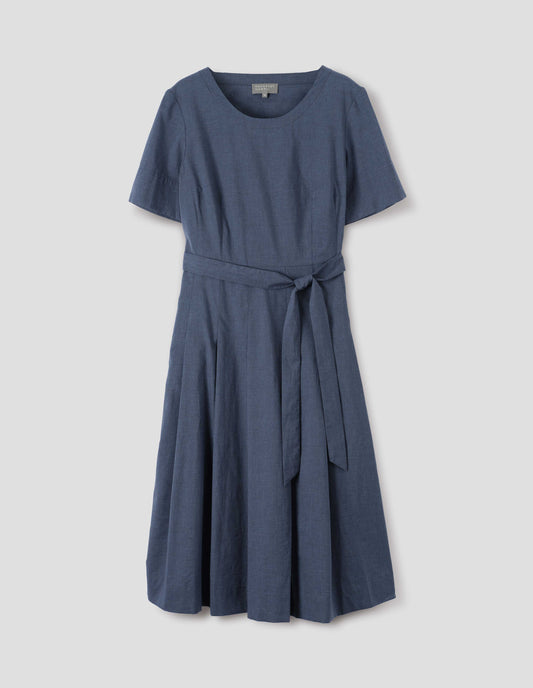 Blue cotton chambray dress, with t-shirt sleeves, deep hem, stitched down pleats, self tie, side pockets and mother of pearl button detailing on one side. Front flat lay view.