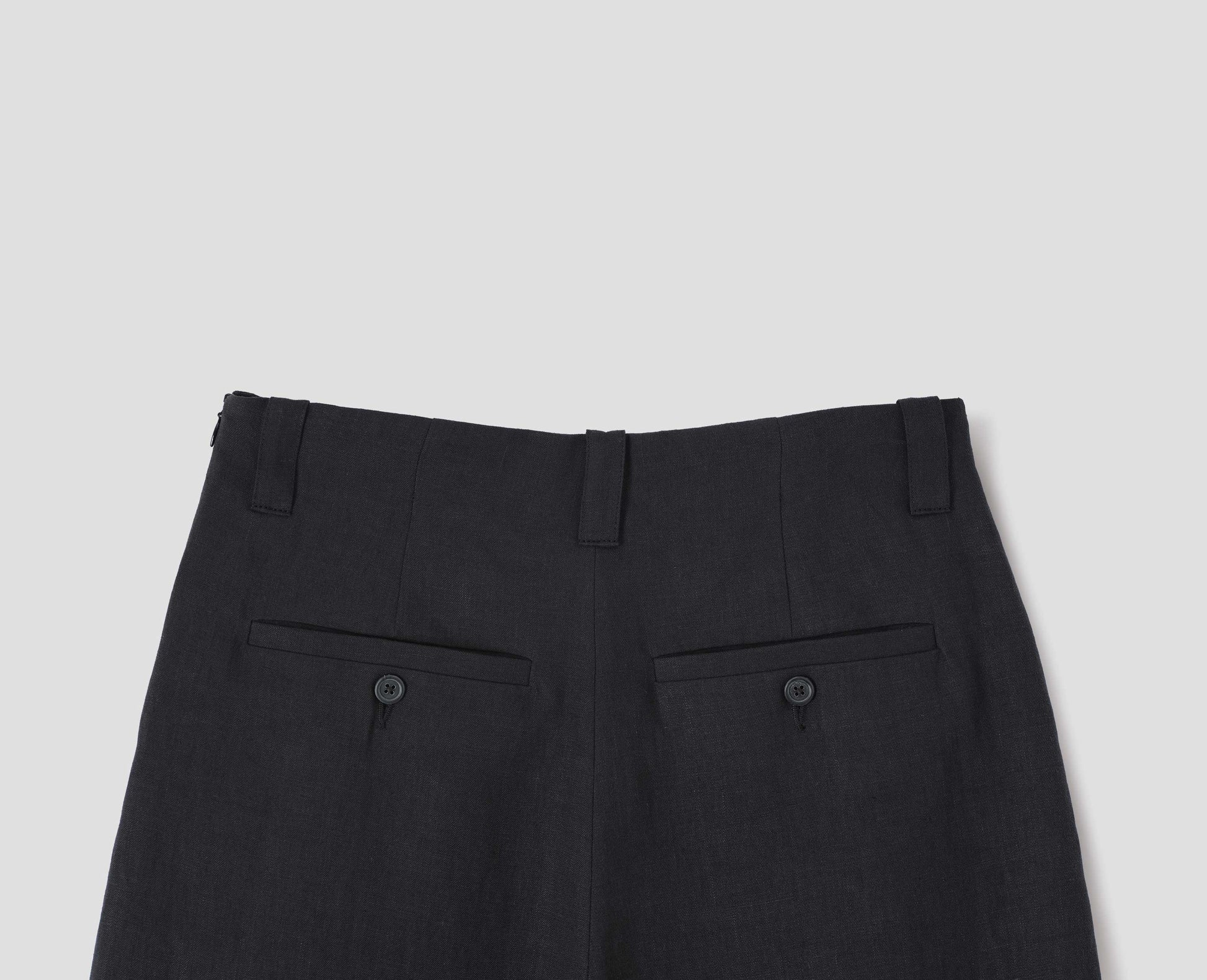 Black culottes with grown-on waistband with belt loops, concealed side zip with horn button tab fastening, and button-through welt back pockets. Back flat lay view.