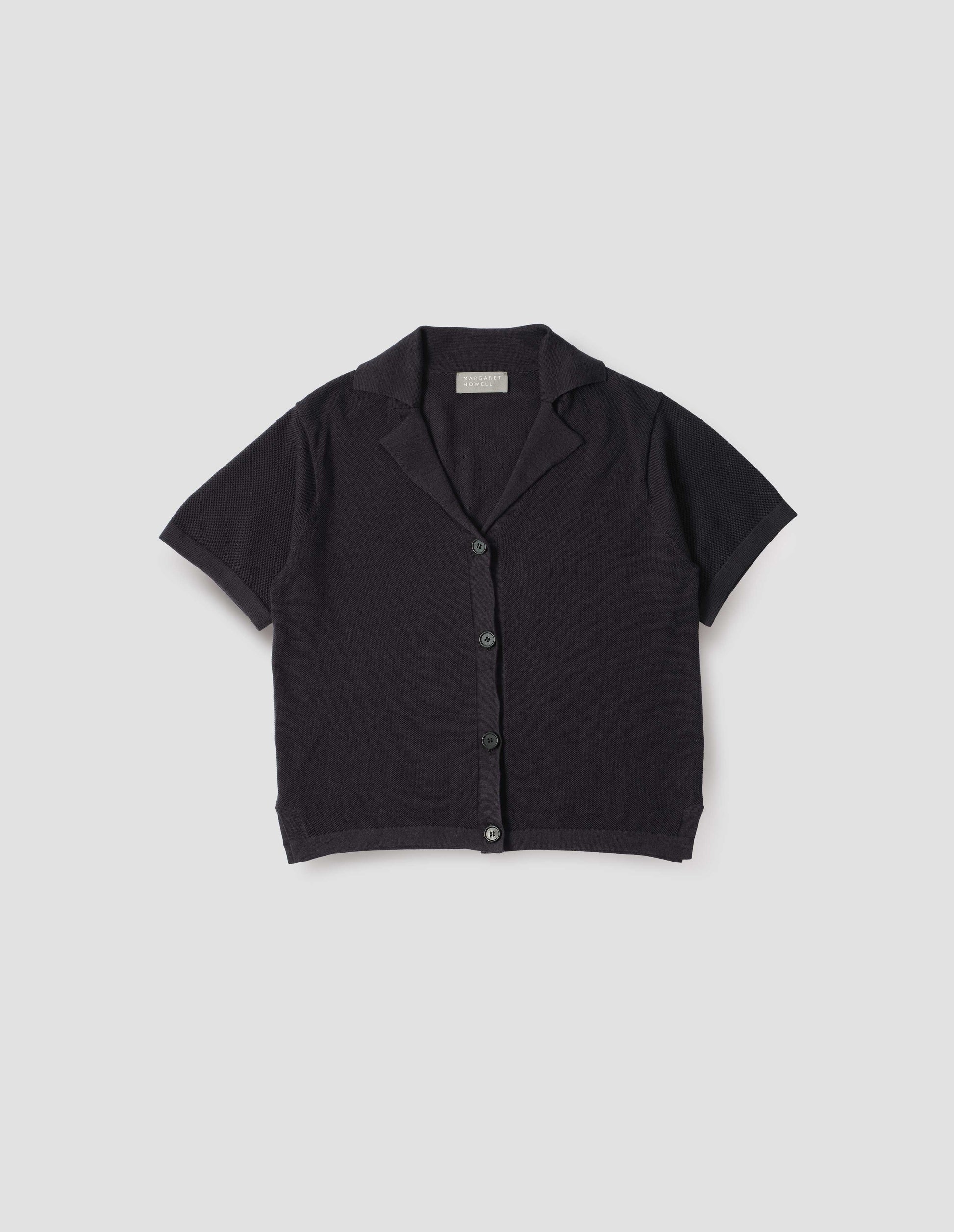 Black knitted cotton short sleeve shirt, with fine gauge rib collar, placket, cuffs and side split hem with horn buttons. Front flat lay view. 