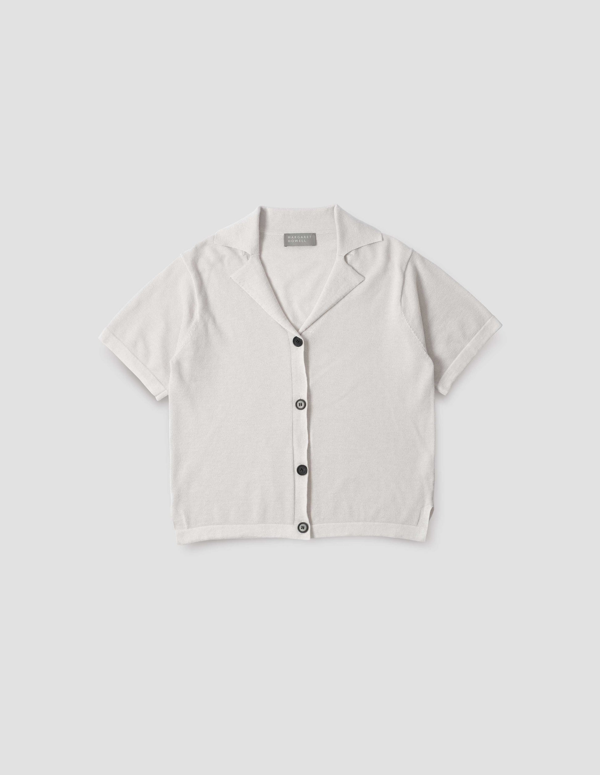 Off white knitted cotton short sleeve shirt, with fine gauge rib collar, placket, cuffs and side split hem with horn buttons. Front flat lay view. 