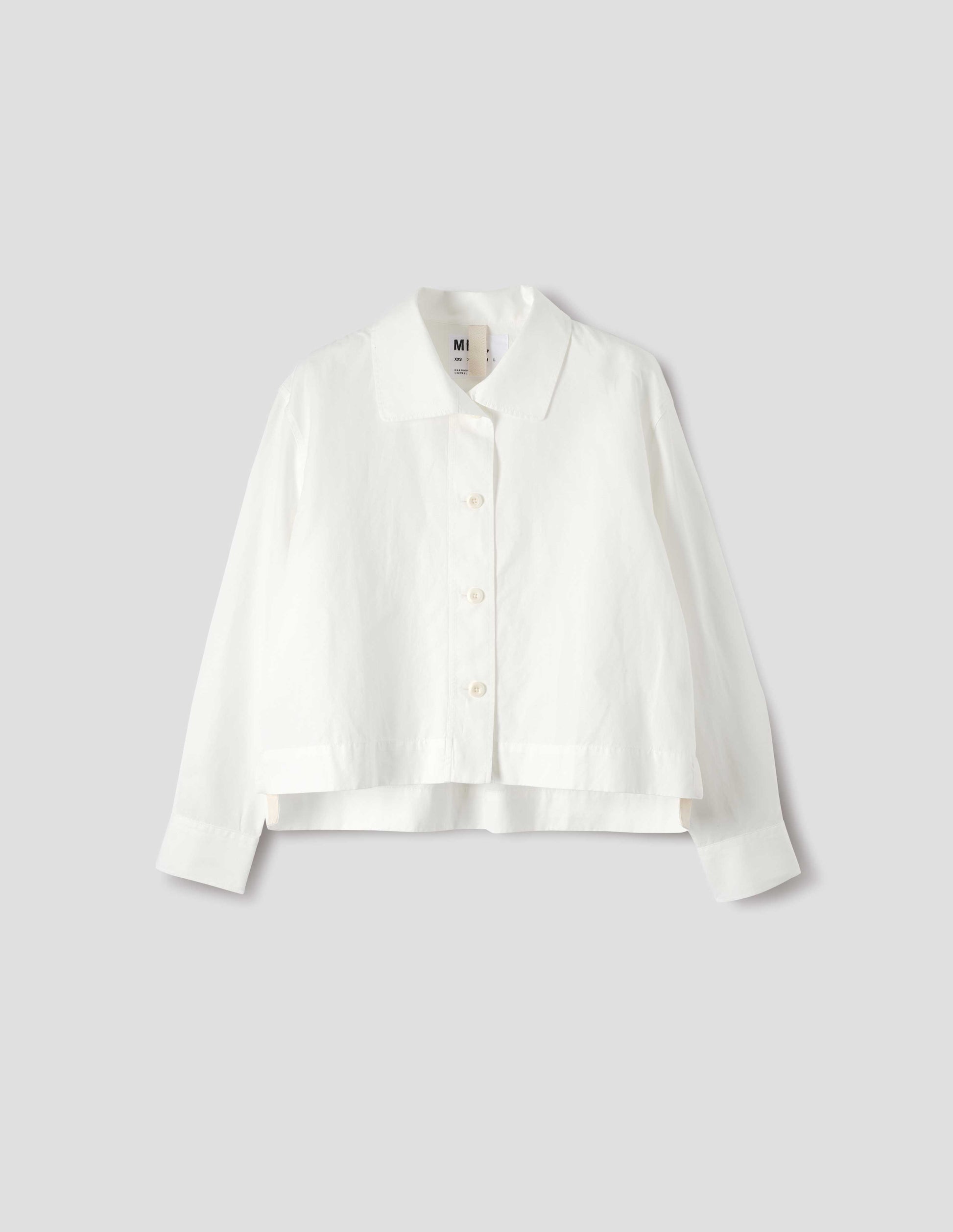 White shirt with round asymmetrical collar, wide stitch-through placket with corozo brace buttons and stepped vent split hem. Front flat lay view.