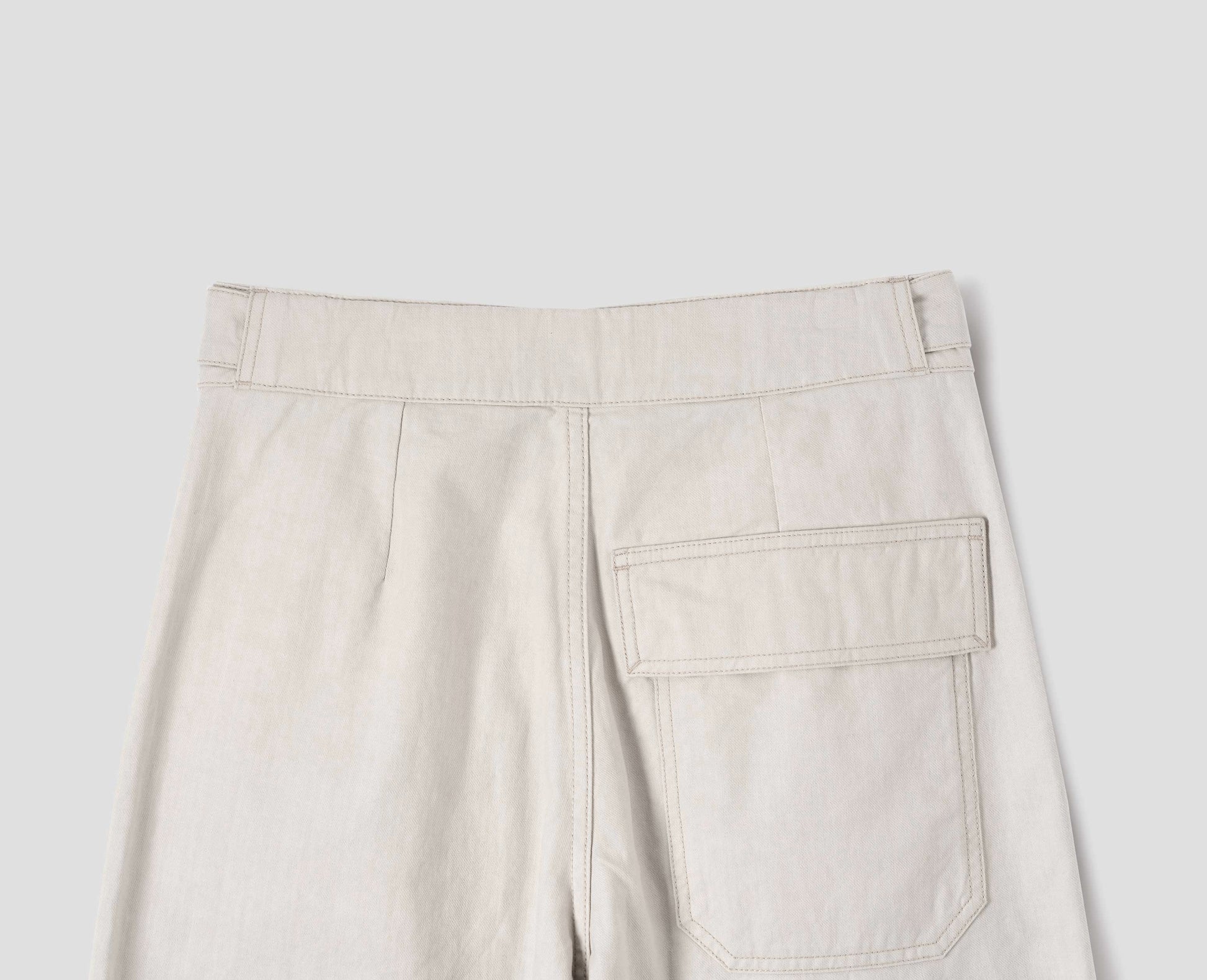 Beige cotton drill trousers with adjustable tab detailing on waistband and flap patch pocket. Back flat lay view.