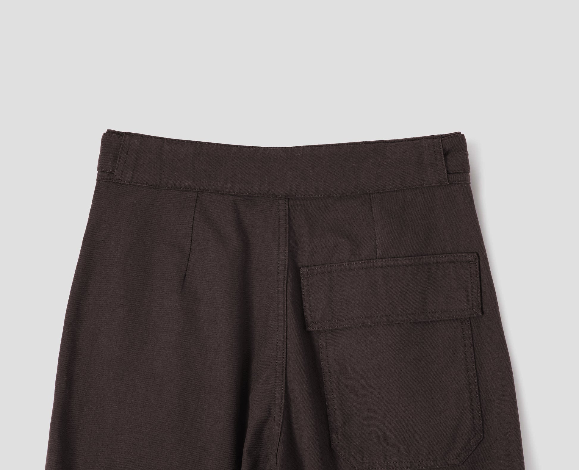 Dark brown cotton drill trousers with adjustable tab detailing on waistband and flap patch pocket. Back flat lay view.