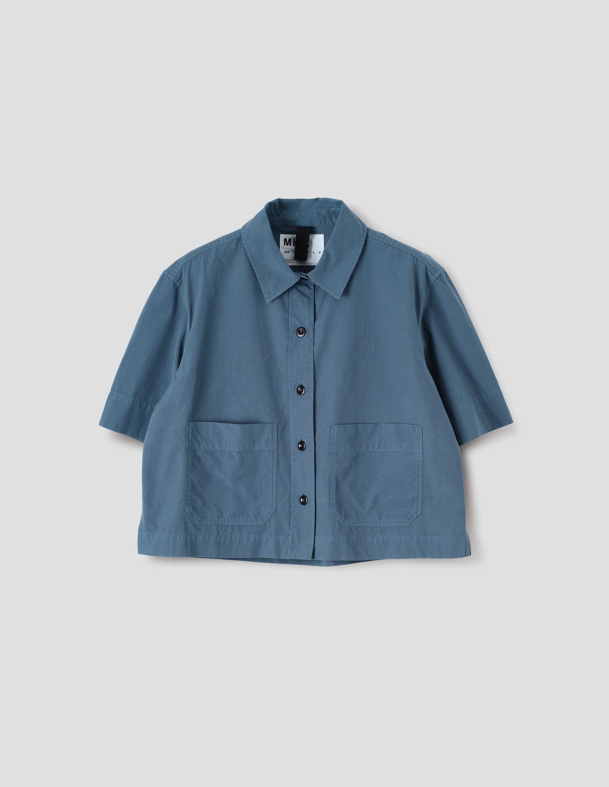 Blue cotton short sleeve shirt, with raised placket, patch pockets, stepped side hem and corozo brace buttons. Front flat lay view. 