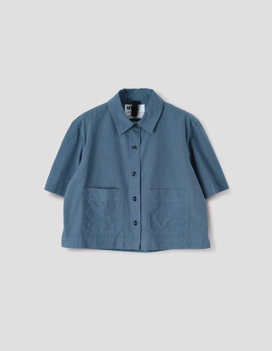 Blue cotton short sleeve shirt, with raised placket, patch pockets, stepped side hem and corozo brace buttons. Front flat lay view. 