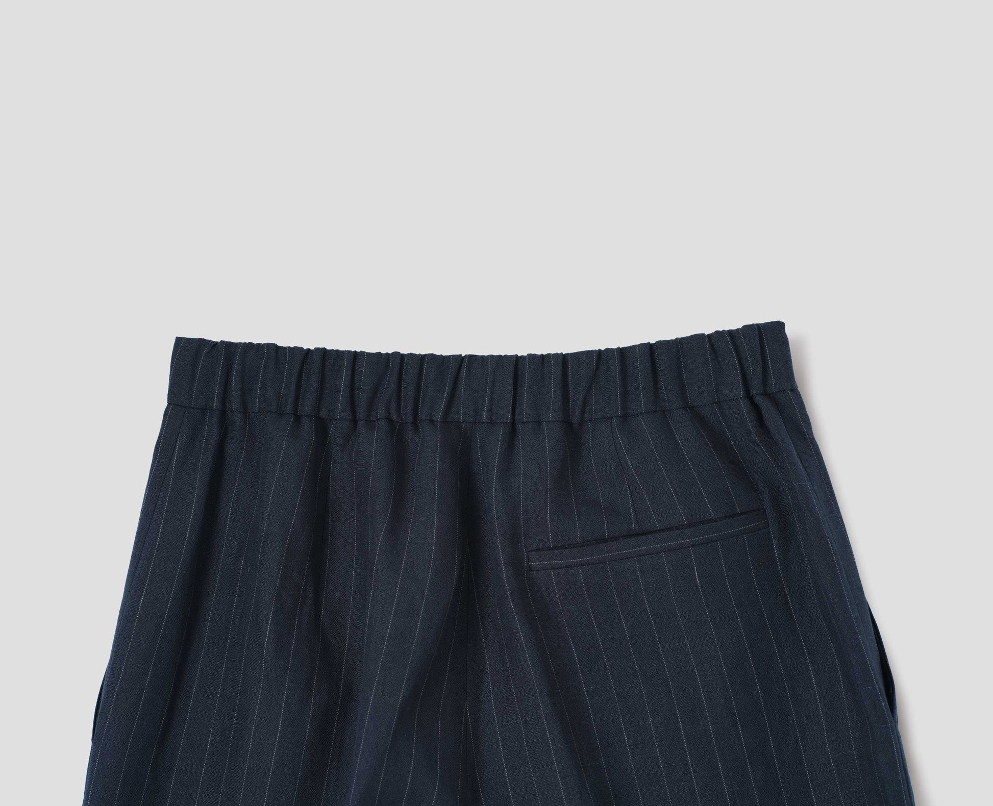 Dark navy and grey pinstripe linen trousers, with half elasticated waistband and belt back pocket. Back flat lay view.